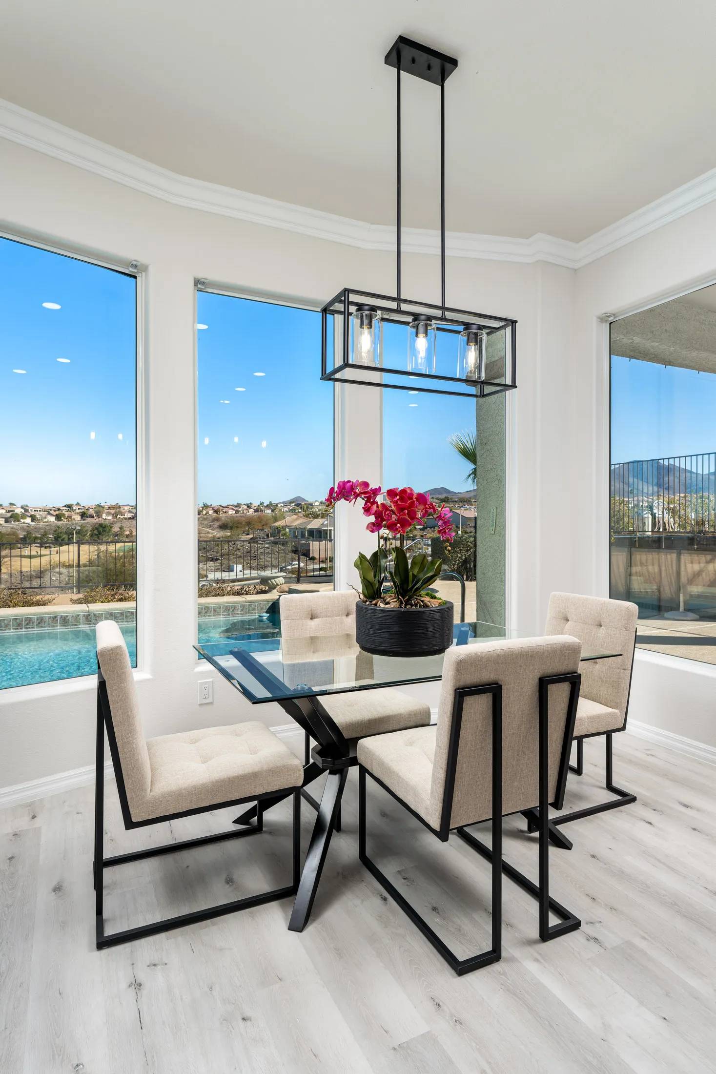 A modern dining nook with a glass-top table, beige upholstered chairs, and large windows offering stunning views of the pool and landscape.