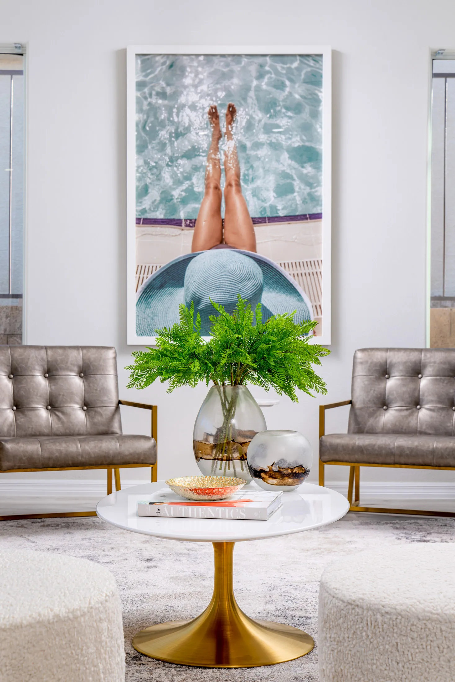 A large photo of legs by a pool hangs above two metallic chairs, with a round gold coffee table and a green plant arrangement in front.