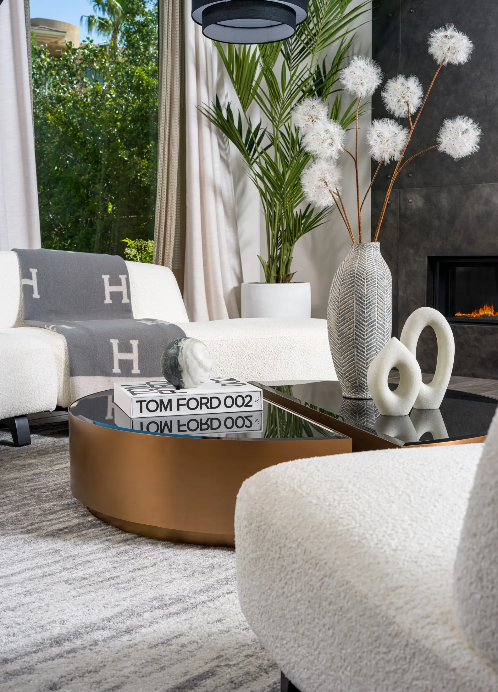 A luxurious living room setup featuring a bronze coffee table with black glass, styled with a designer book, marble accent, textured vase, and sculptural decor. The room includes a white curved sofa, lush greenery, and a sleek fireplace, showcasing Stately’s luxury home staging expertise in Las Vegas.
