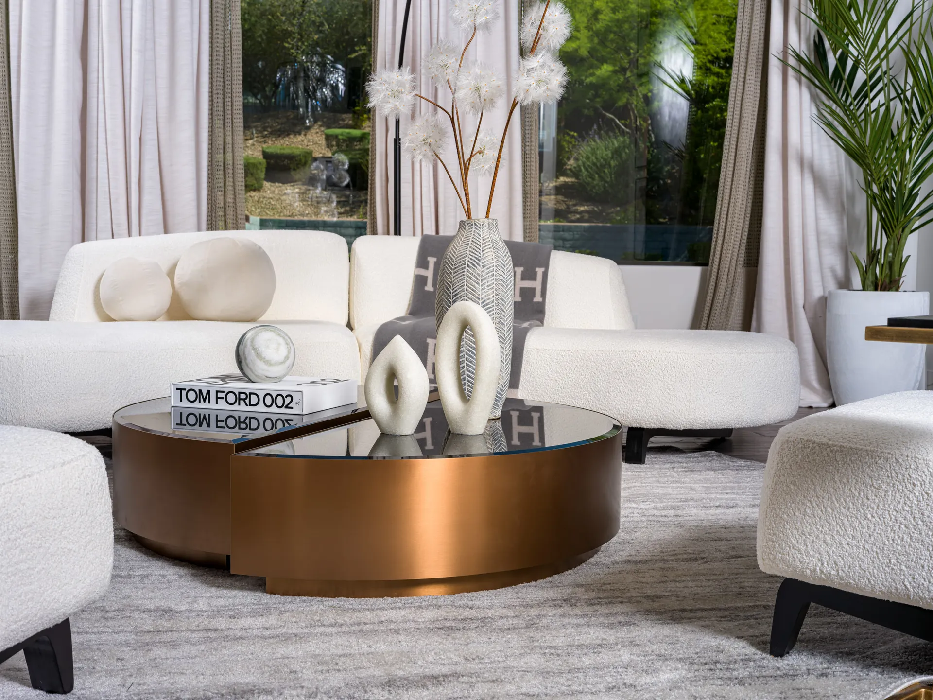 A modern living room featuring a bronze coffee table with black glass, styled with sculptural decor and designer books, paired with a curved white sofa and floor-to-ceiling windows overlooking lush greenery. This setup highlights Stately’s luxury home staging expertise in Las Vegas.