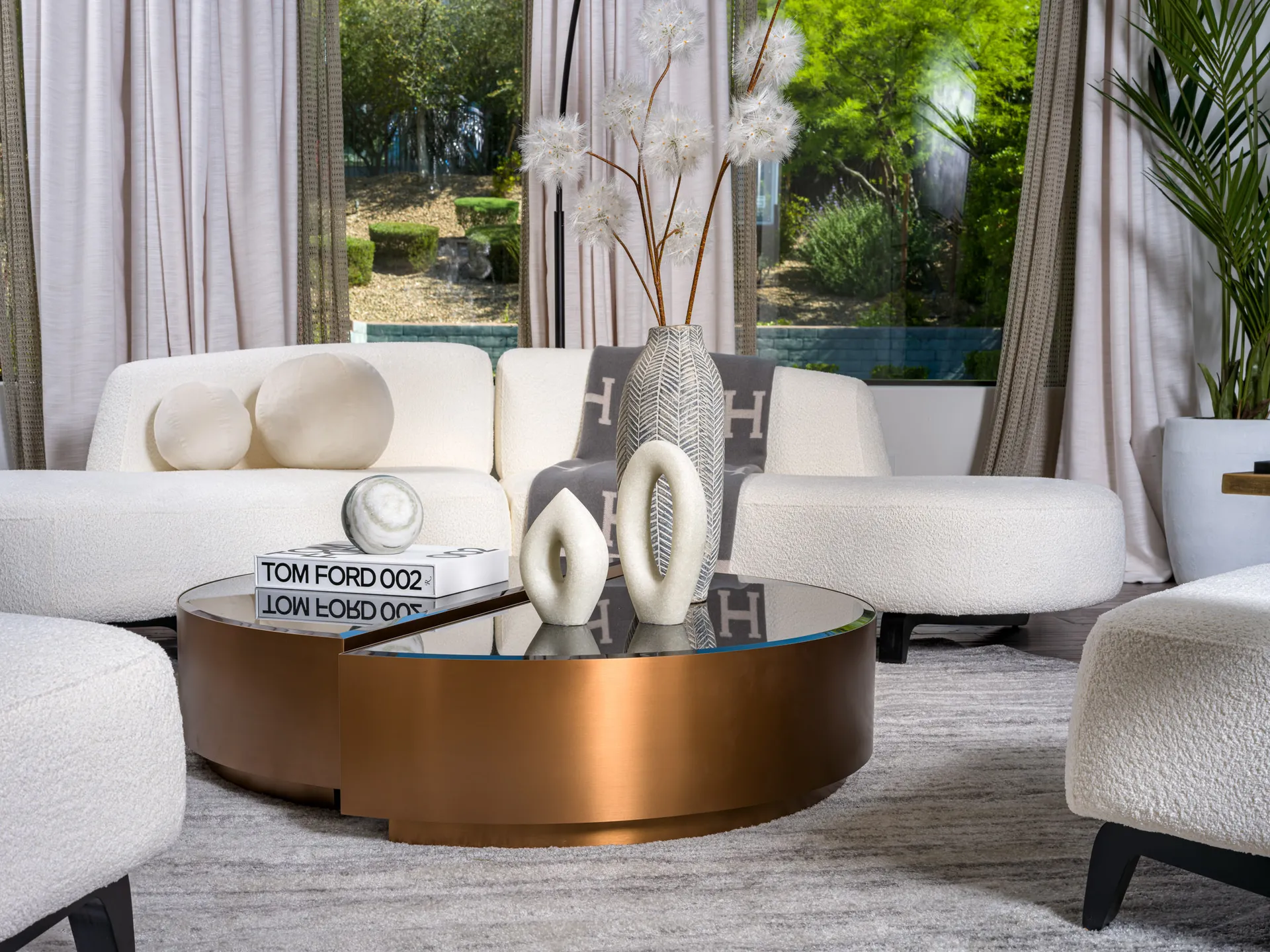 A contemporary living room setup featuring a bronze and black glass coffee table with sculptural decor, a designer book, and a cozy white curved sofa. Floor-to-ceiling windows offer lush outdoor views, exemplifying Stately’s expertise in luxury home staging and interior design in Las Vegas.