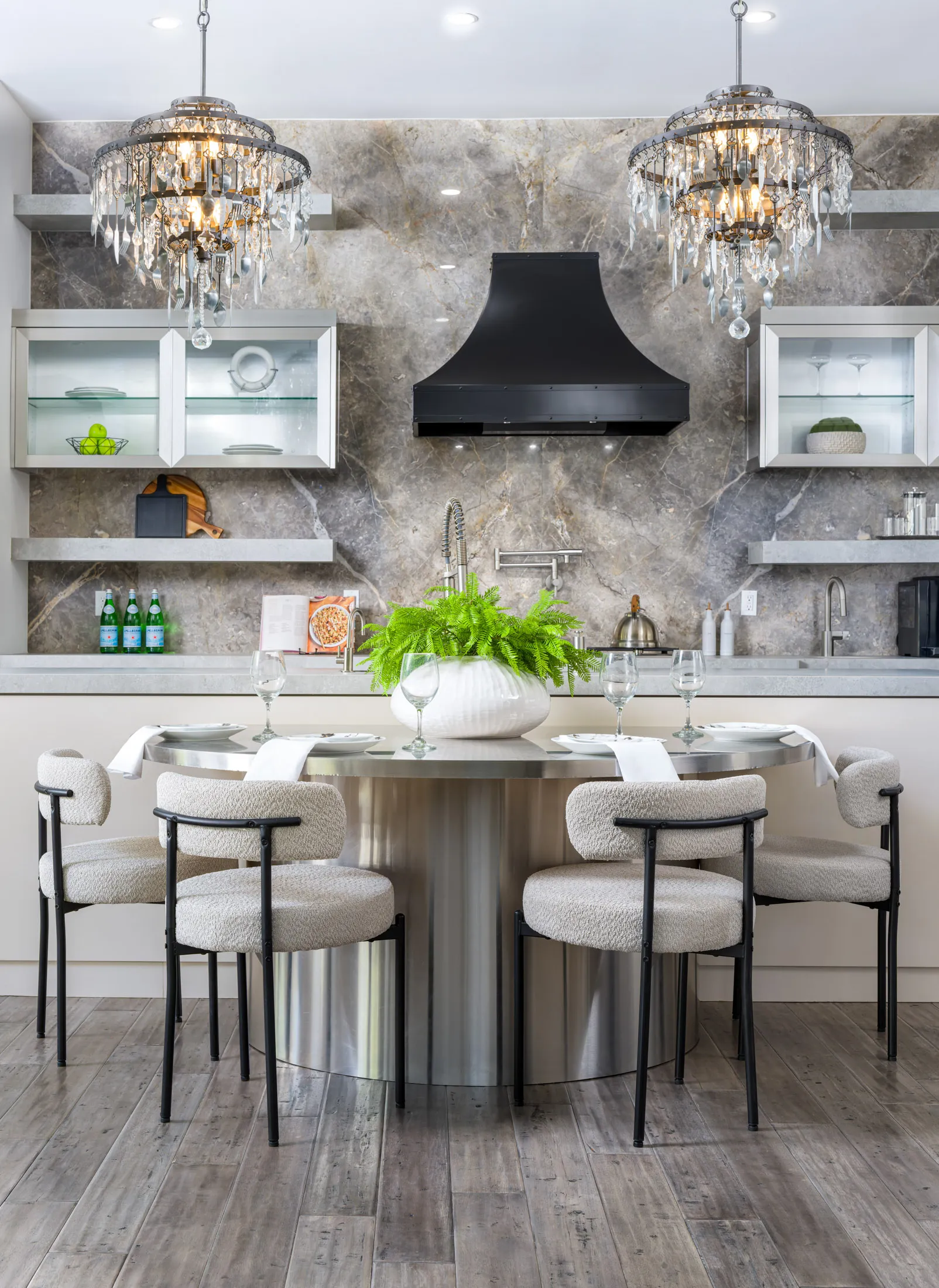 A luxurious kitchen and dining area featuring a round stainless steel table, modern upholstered chairs, a marble backsplash, a black range hood, and sparkling chandeliers. This design highlights Stately’s expertise in creating sophisticated spaces through luxury home staging in Las Vegas.