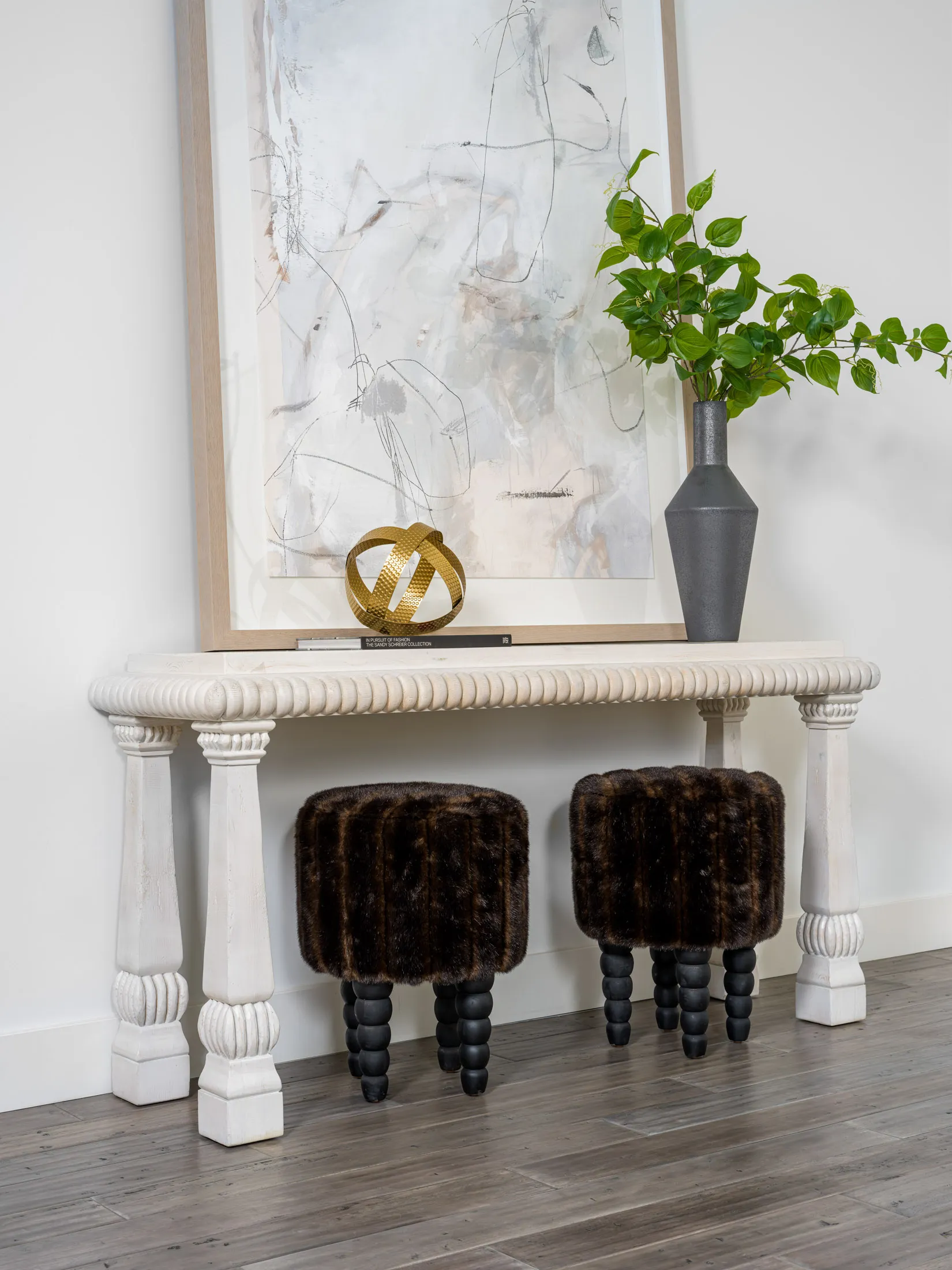 A carved white console table topped with an abstract painting, a gold accent sculpture, and a tall vase of green leaves. Two plush, fur-like stools sit neatly beneath.