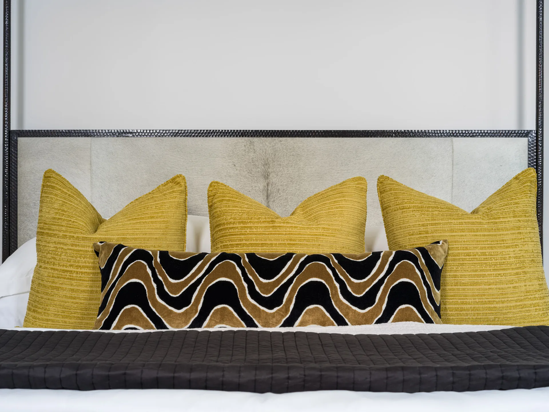 Close-up of a black-framed headboard with bright mustard and patterned accent pillows. The pillows feature rich textures and a wavy design.