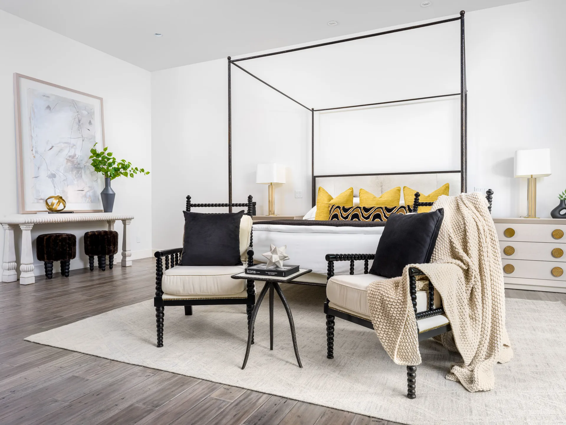 A black canopy bed with bold yellow accent pillows, flanked by two spindle chairs. An abstract painting rests on a console table, and dark wood flooring ties the space together.