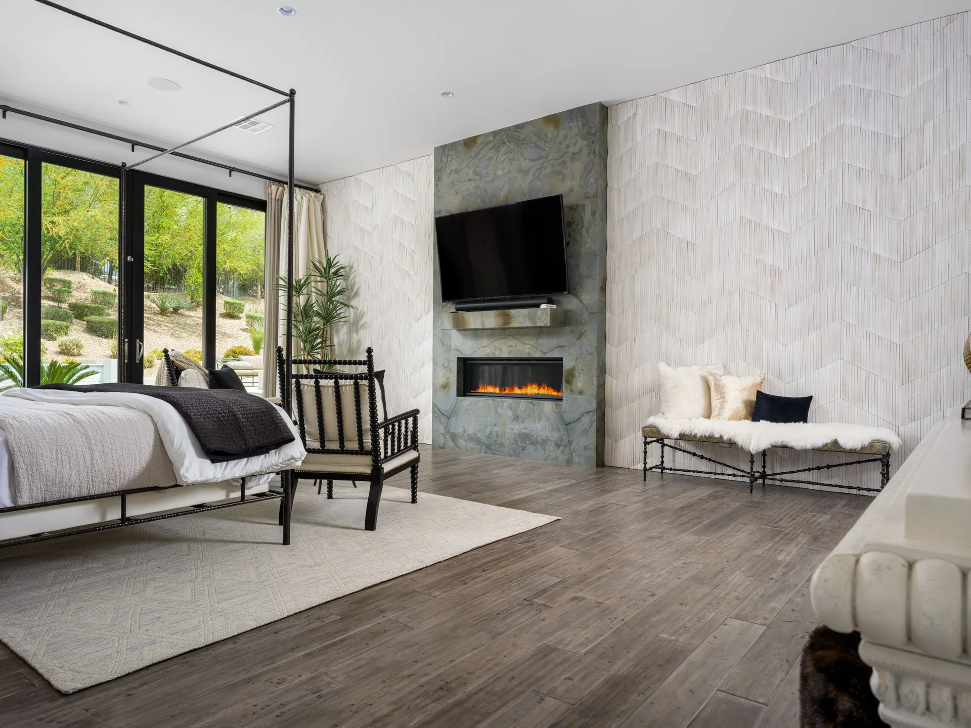 A bedroom featuring a black canopy bed, a stone fireplace wall, and large windows overlooking landscaped greenery. A plush bench sits along a textured accent wall.