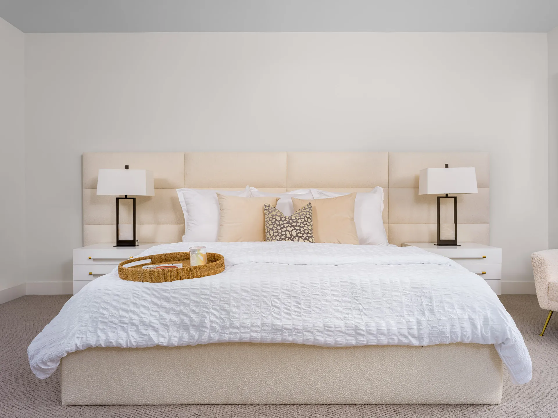 A large bed with an upholstered headboard, white bedding, and neutral pillows. Matching white nightstands with black lamps flank the bed.