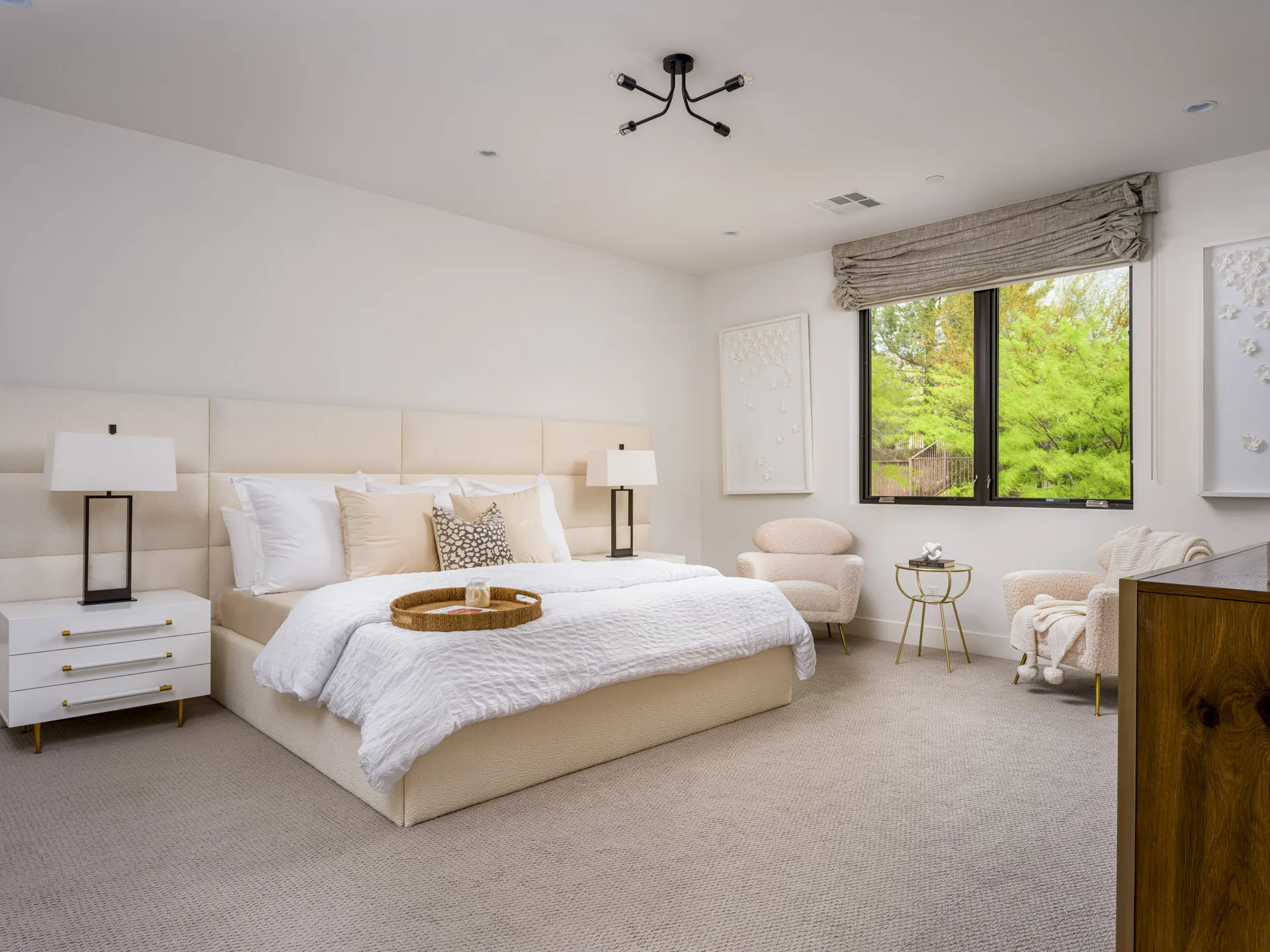 A bright bedroom featuring a large upholstered headboard, white bedding, and two cozy chairs by the window. Neutral tones and soft textures create a serene atmosphere.