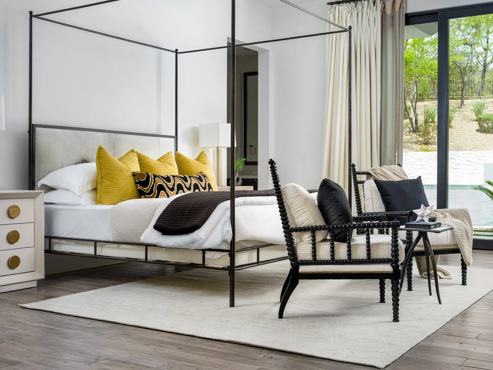 A luxurioLuxurious bedroom with modern canopy bed, yellow pillows, and outdoor viewus bedroom at Lookout Ridge Dr. featuring a modern canopy bed with vibrant yellow and patterned accent pillows, a sleek black spindle chair with neutral cushions, and floor-to-ceiling windows overlooking a serene outdoor space.
