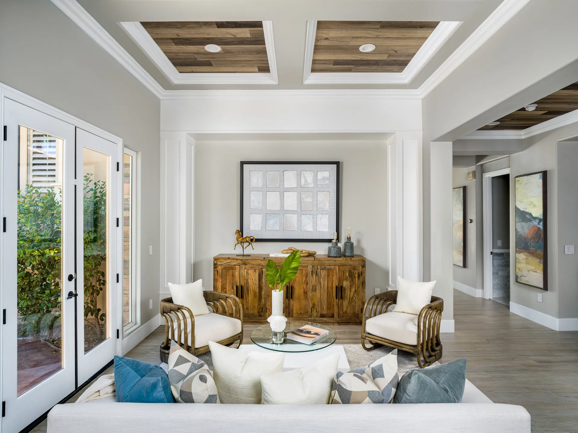 A living room with a wooden console, rattan chairs, and coffered ceilings, featuring glass doors and neutral furnishings.