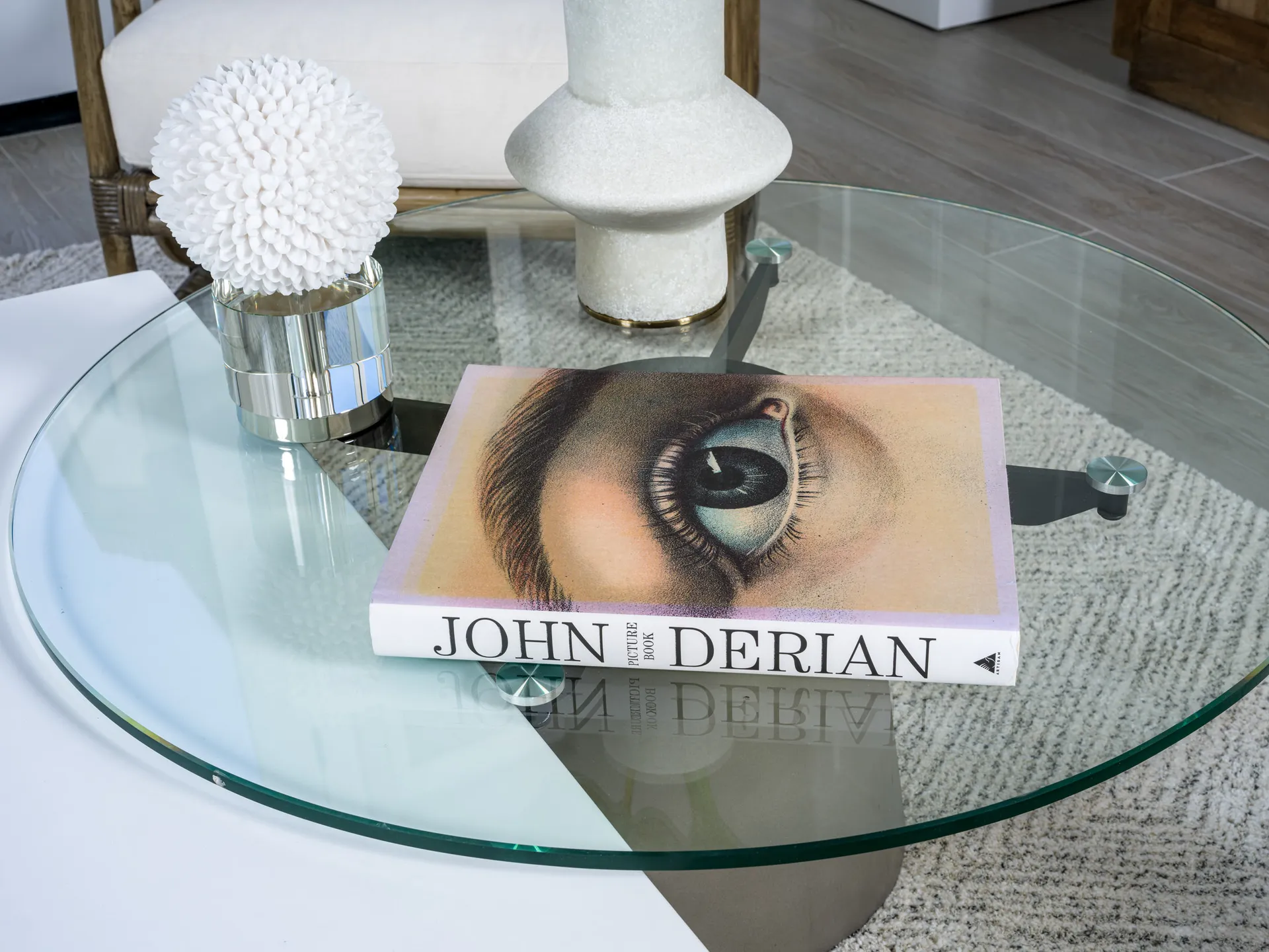 A round glass coffee table featuring a John Derian book with an eye illustration and a white decorative sphere.