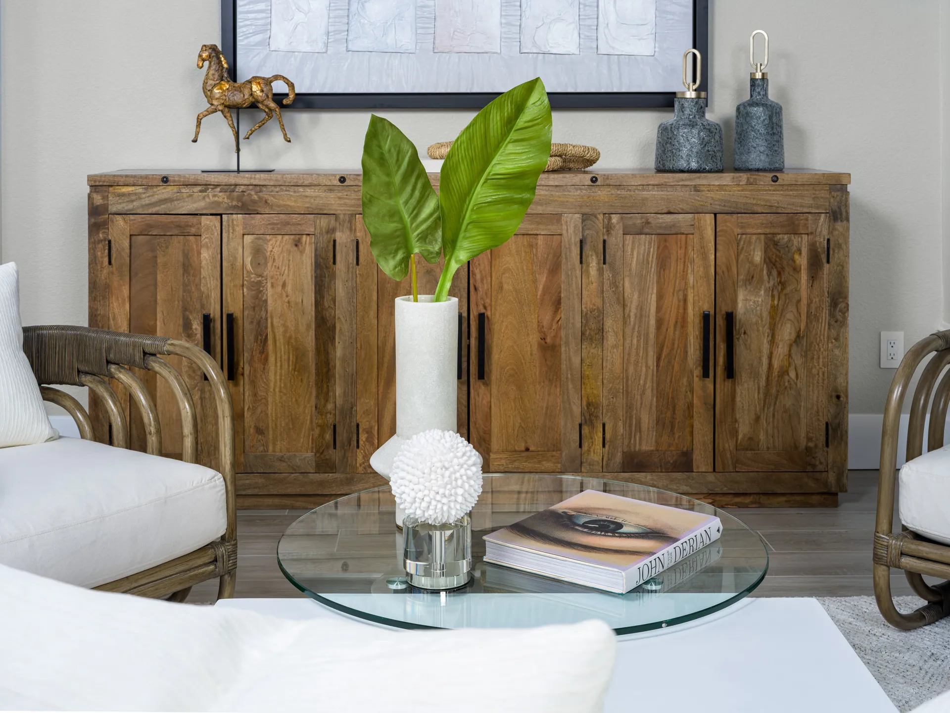 A living room, styled by Modern Rustic Home Staging, featuring a wooden console, a gold horse sculpture, and a tall green leaf in a white vase on a glass coffee table, surrounded by rattan chairs.
