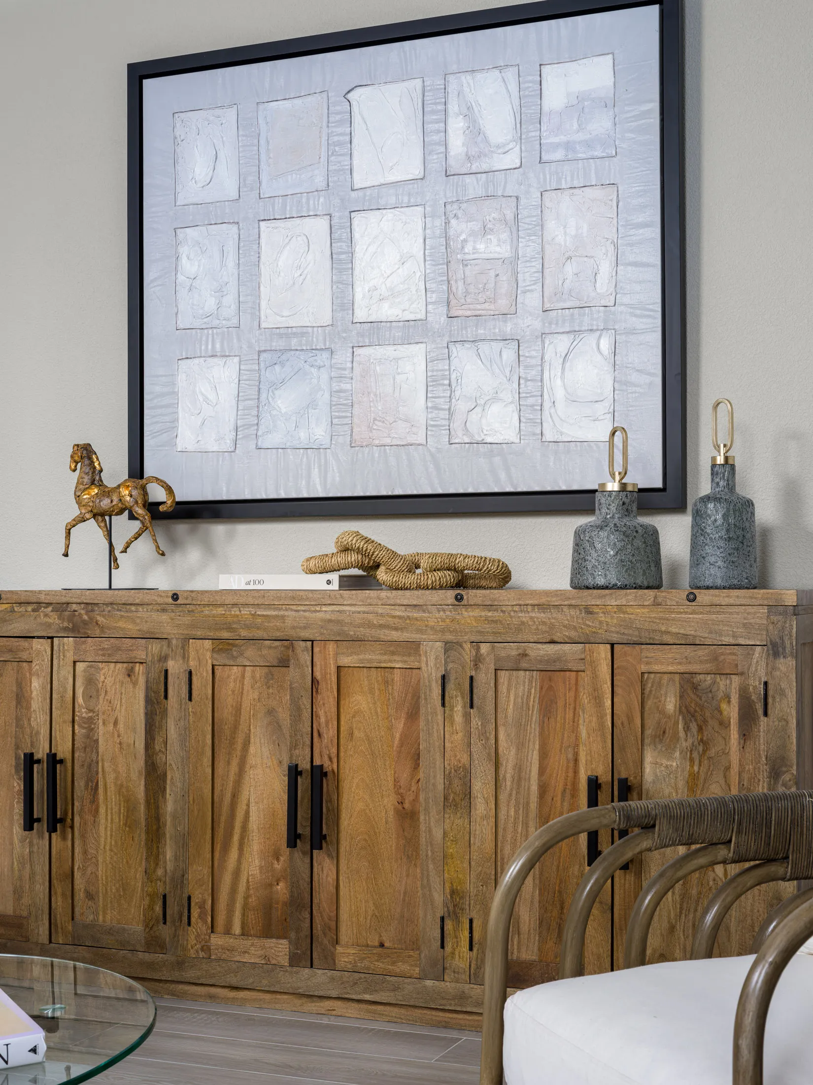 A wooden console with black metal handles topped by a gold horse figurine, rope accents, and two gray vases under a grid-style abstract painting.