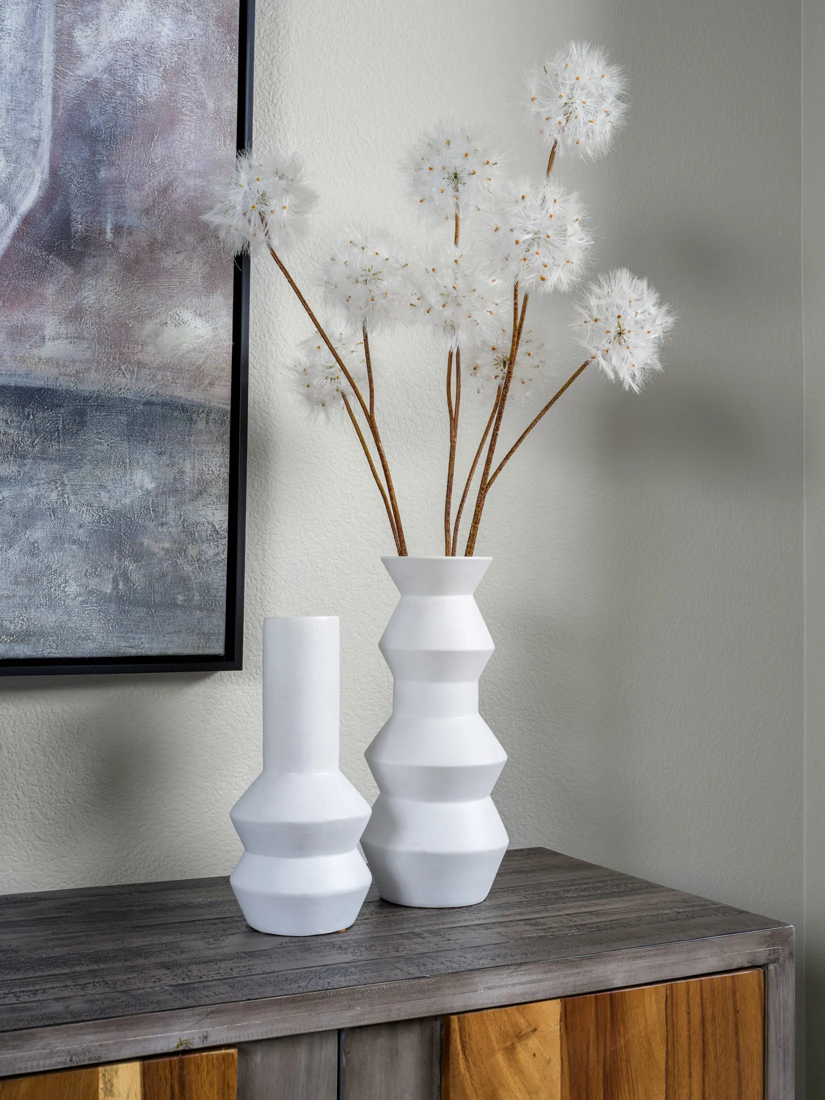 A pair of white geometric vases on a wooden console table, featuring delicate dandelion-like stems.