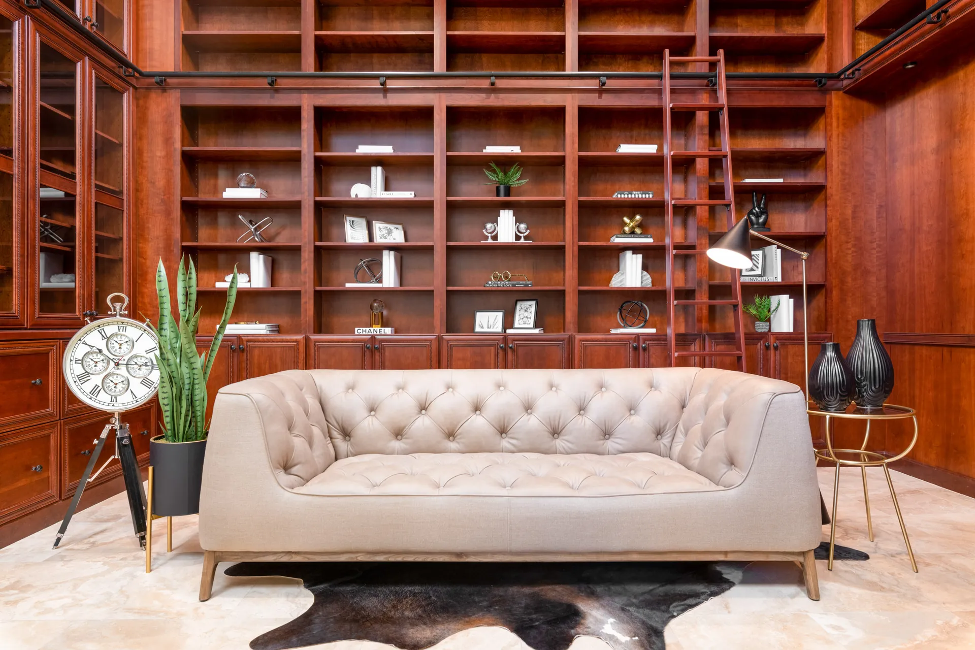 Luxury Home Staging A tufted beige sofa set against built-in wooden shelves with a rolling ladder. A large standing clock and a snake plant add visual interest.