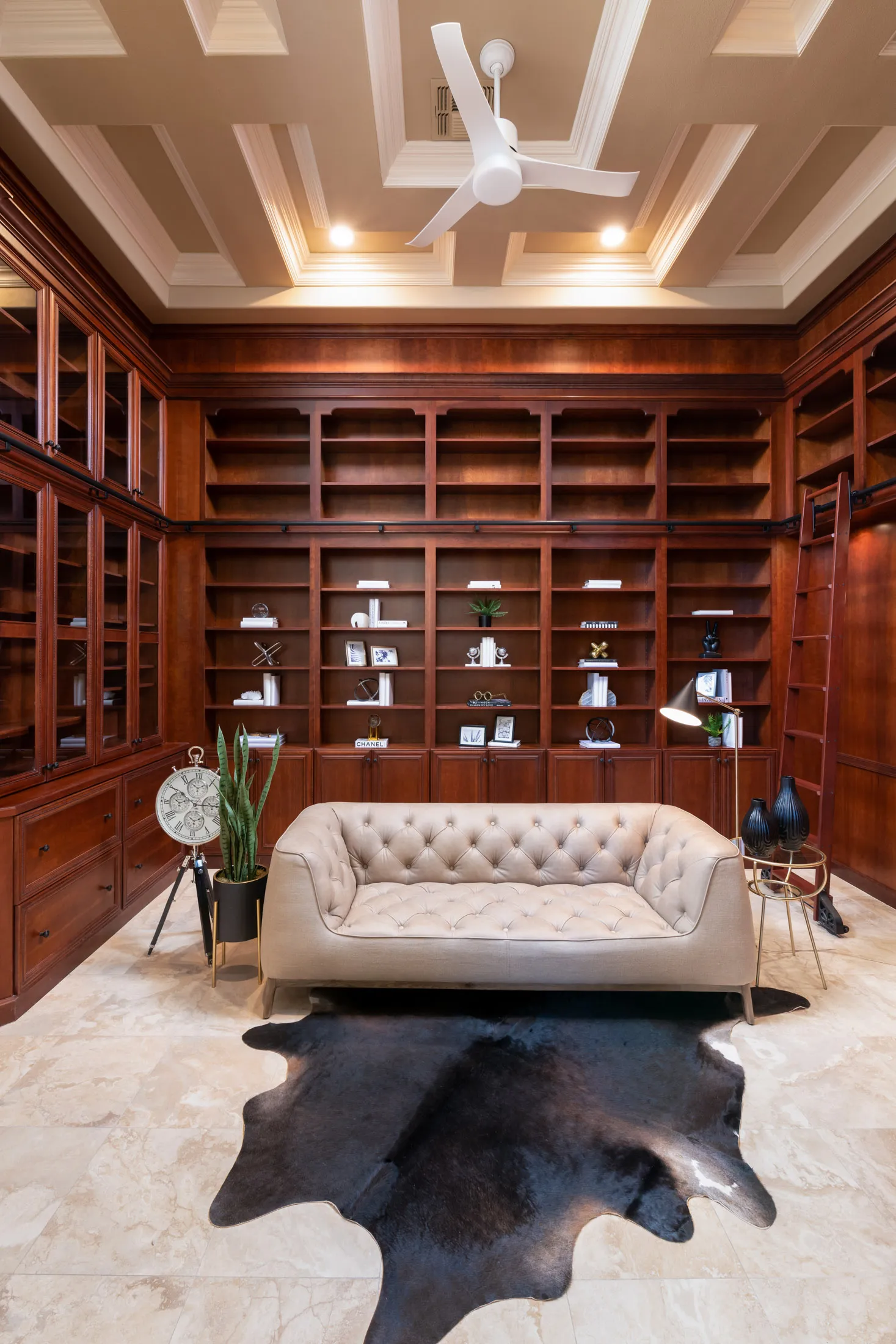 A large library with built-in wooden shelves, a tufted sofa, and a black hide rug under a coffered ceiling.