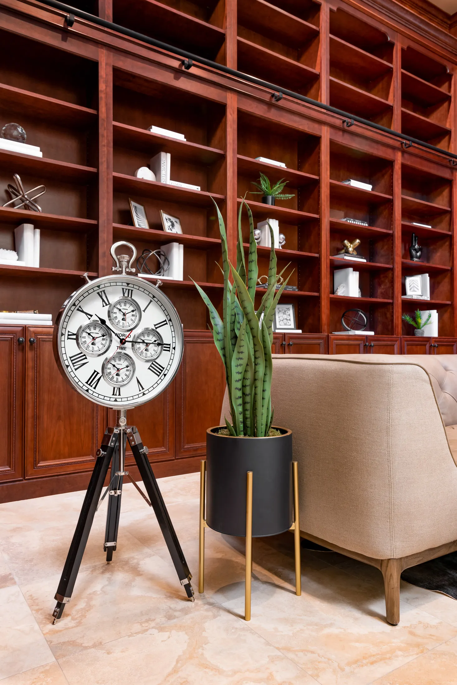 A tall, vintage-style clock on a tripod stand next to a snake plant in a black planter, set before cherry wood bookshelves filled with decorative accents.