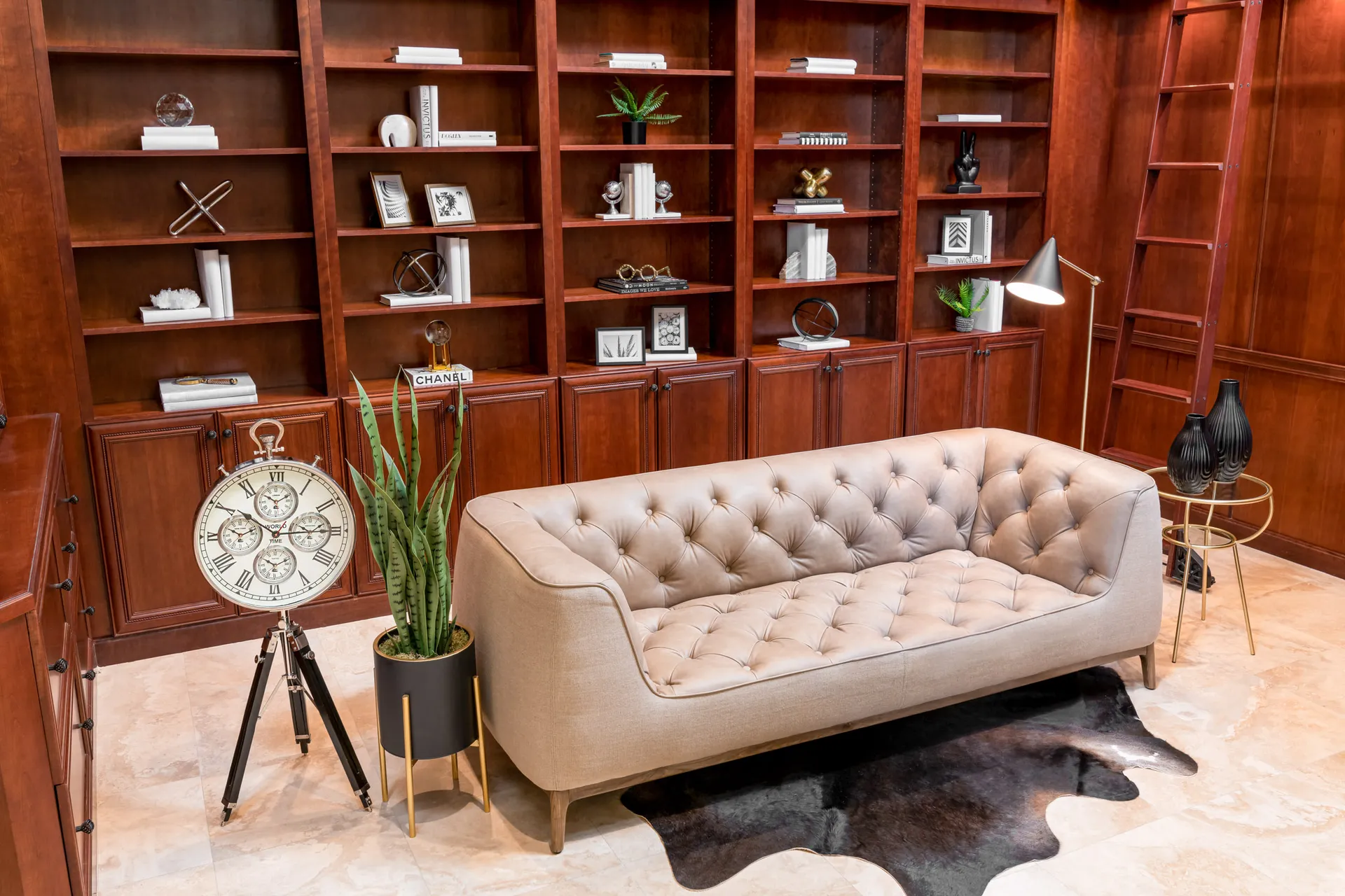 A tufted sofa in front of tall, built-in wooden shelves adorned with decorative objects. A large standing clock and a potted snake plant sit nearby.