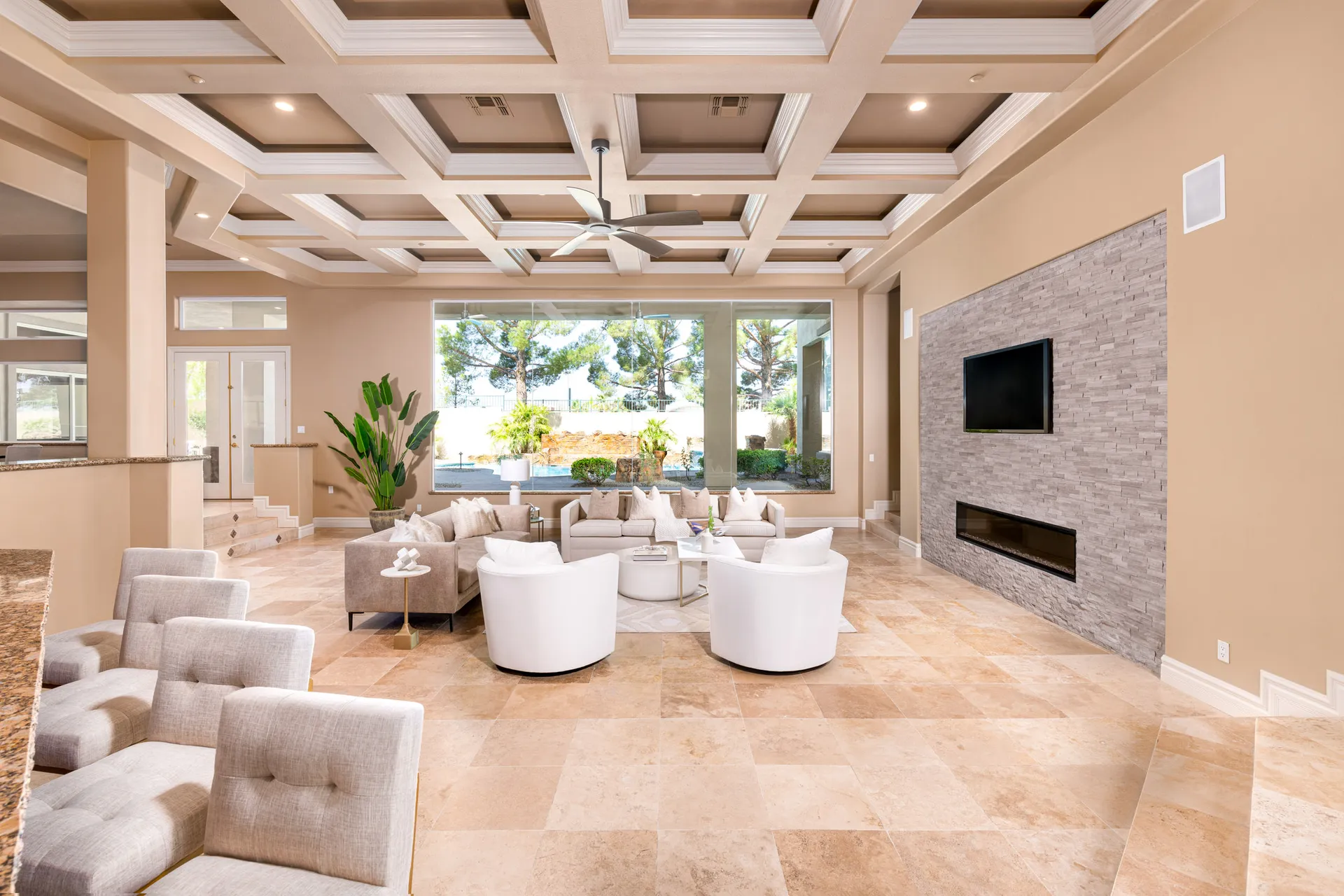 A spacious living room with a coffered ceiling, large windows, neutral seating, and a sleek stone fireplace wall.