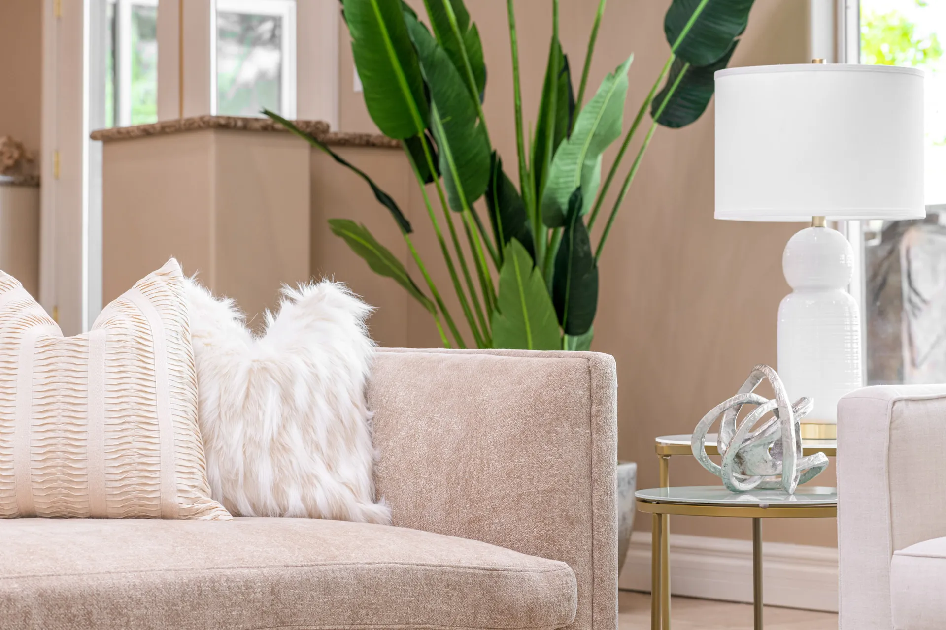 A beige sofa adorned with a striped pillow and a fluffy white pillow, next to a white table lamp and tall green foliage.