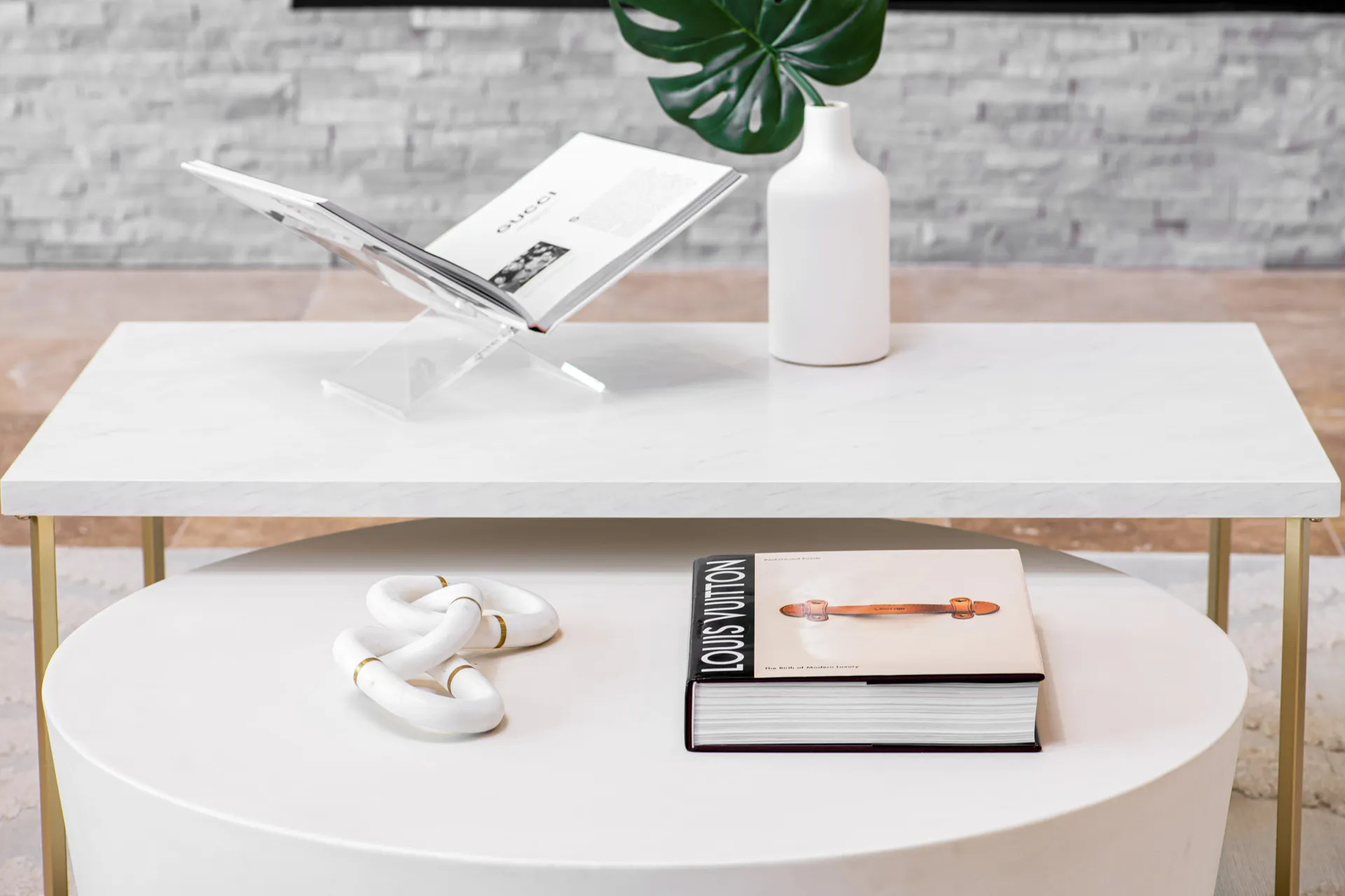 A two-tier white coffee table displaying an open designer book on a stand, a potted green leaf, and a decorative white chain accent.