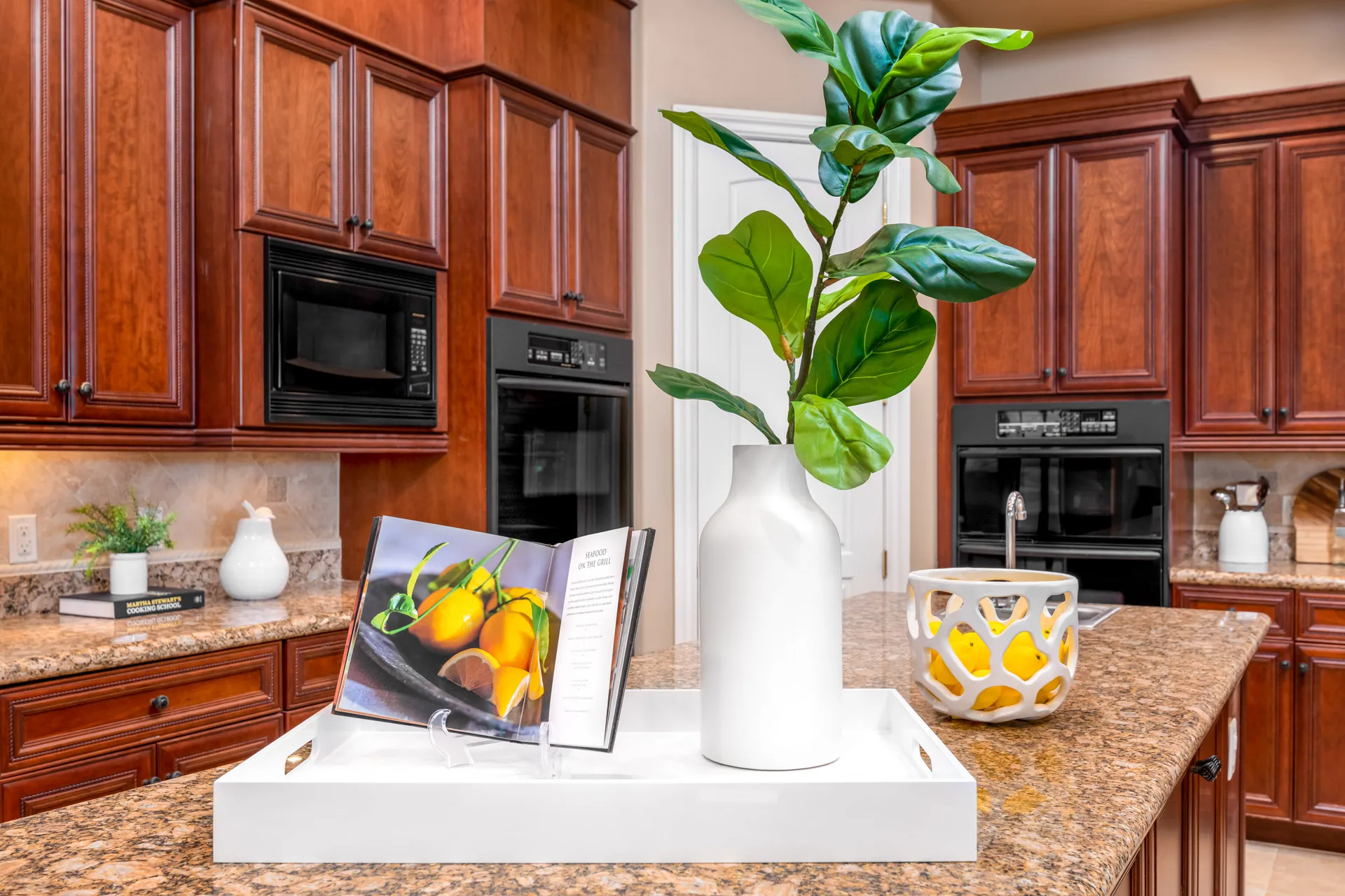 A white tray on a granite island holding a tall green plant, an open cookbook, and a decorative bowl of lemons.