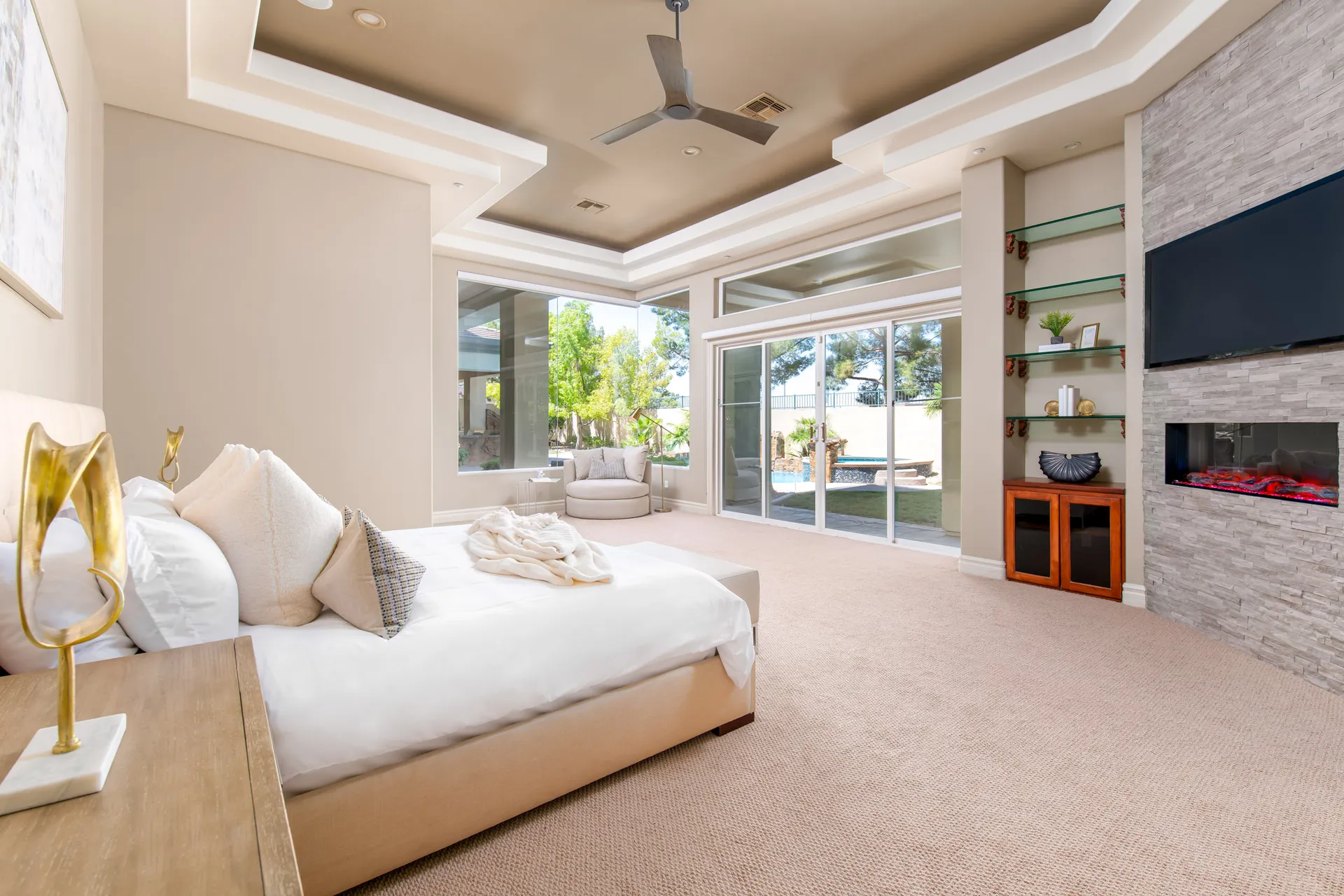 A spacious master bedroom with a neutral bed, modern fireplace, built-in shelves, and sliding doors leading to the backyard pool.