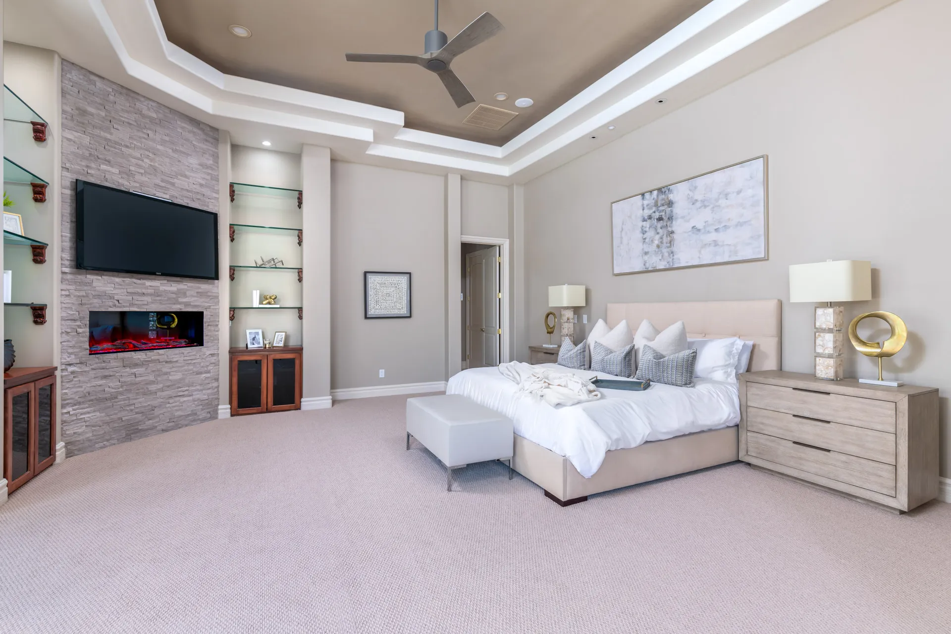 A spacious, neutral-toned bedroom featuring a built-in electric fireplace and TV surrounded by glass shelving.