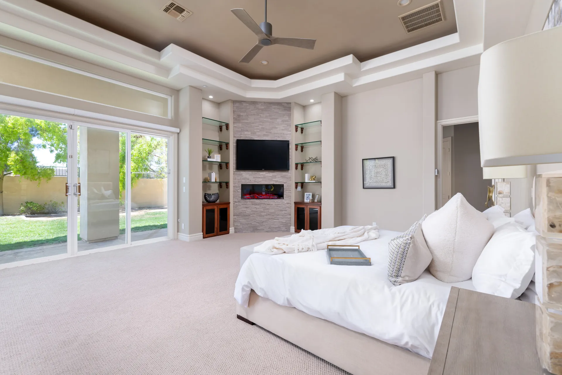 A neutral master bedroom featuring built-in shelves, a fireplace with a TV above, and sliding glass doors opening to a backyard.