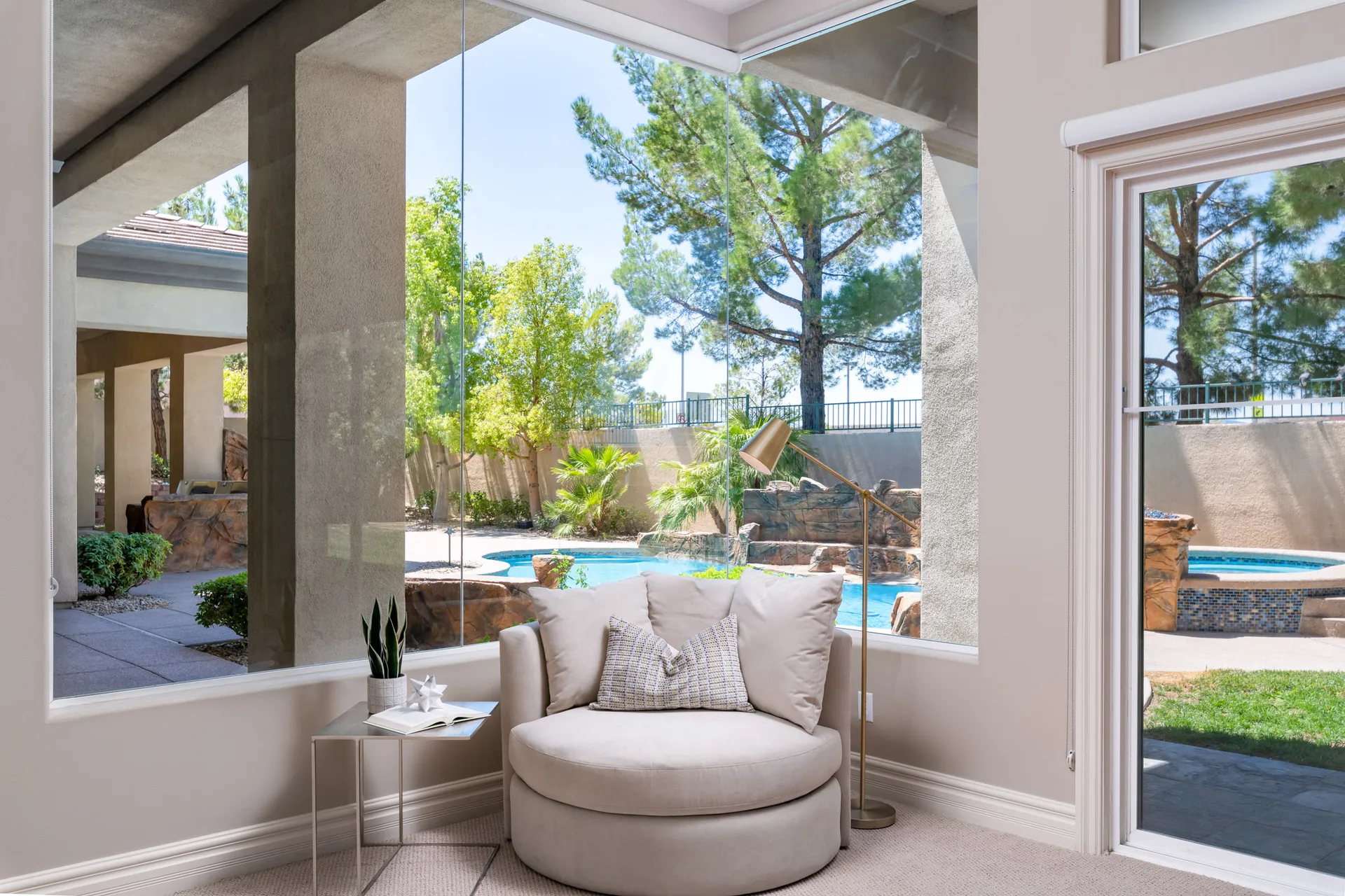 A round upholstered chair near a large window with views of a backyard pool and greenery.
