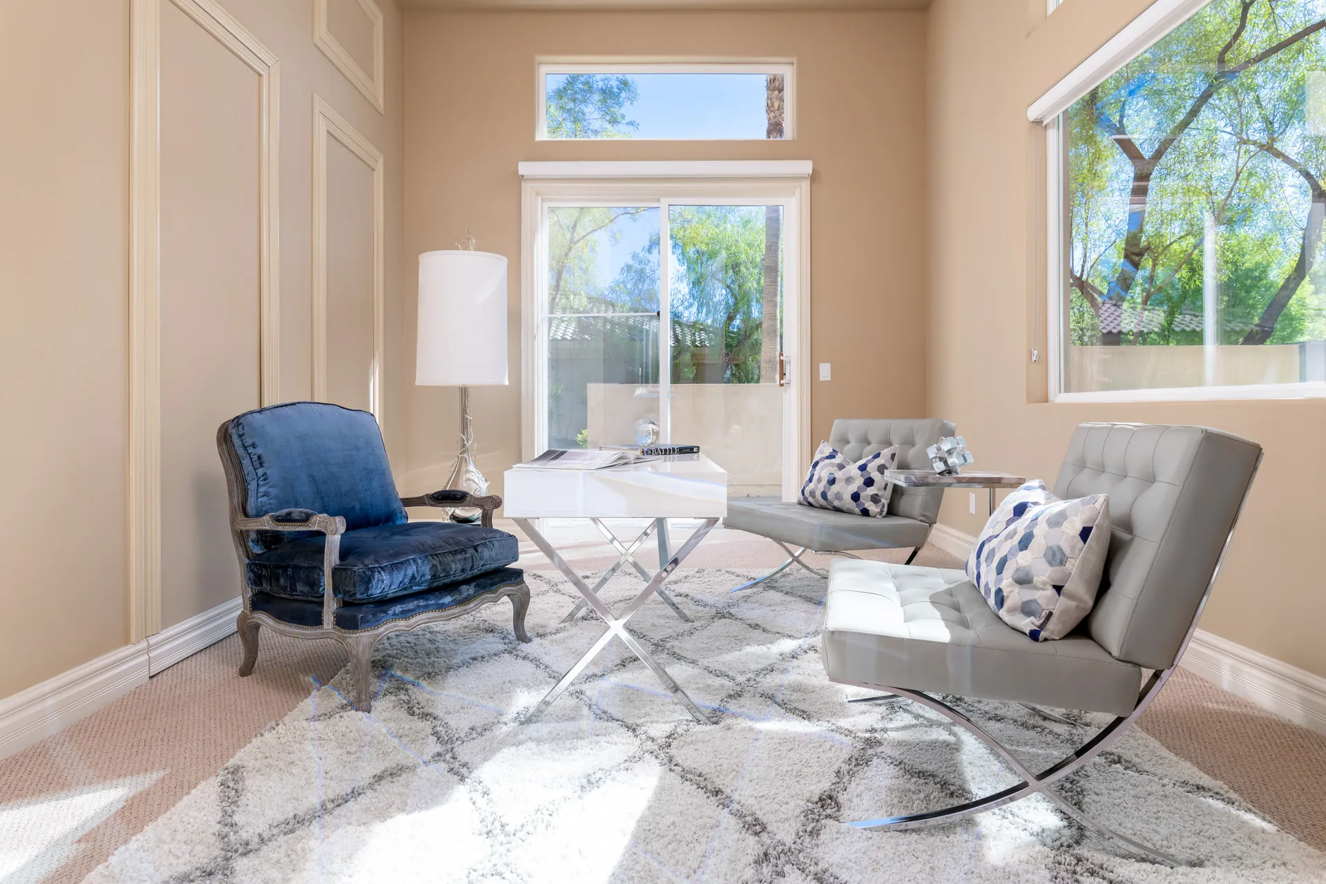 A bright sitting area featuring a navy velvet armchair, tufted gray chairs, a white desk, and large windows revealing an outdoor view.