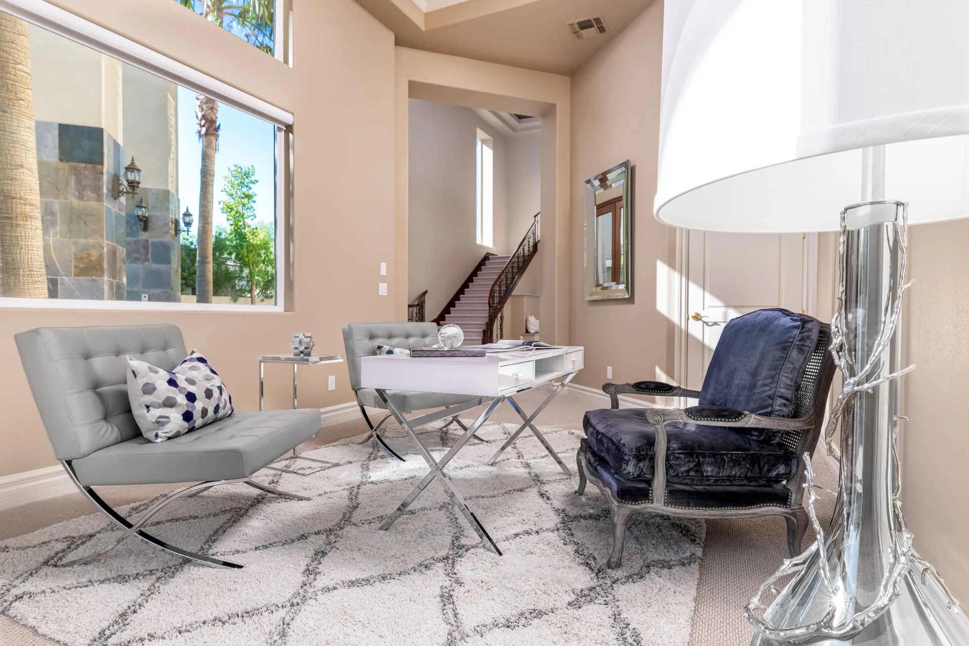 A bright sitting area featuring tufted gray chairs, a navy velvet armchair, a white desk, and large windows overlooking the outdoors.