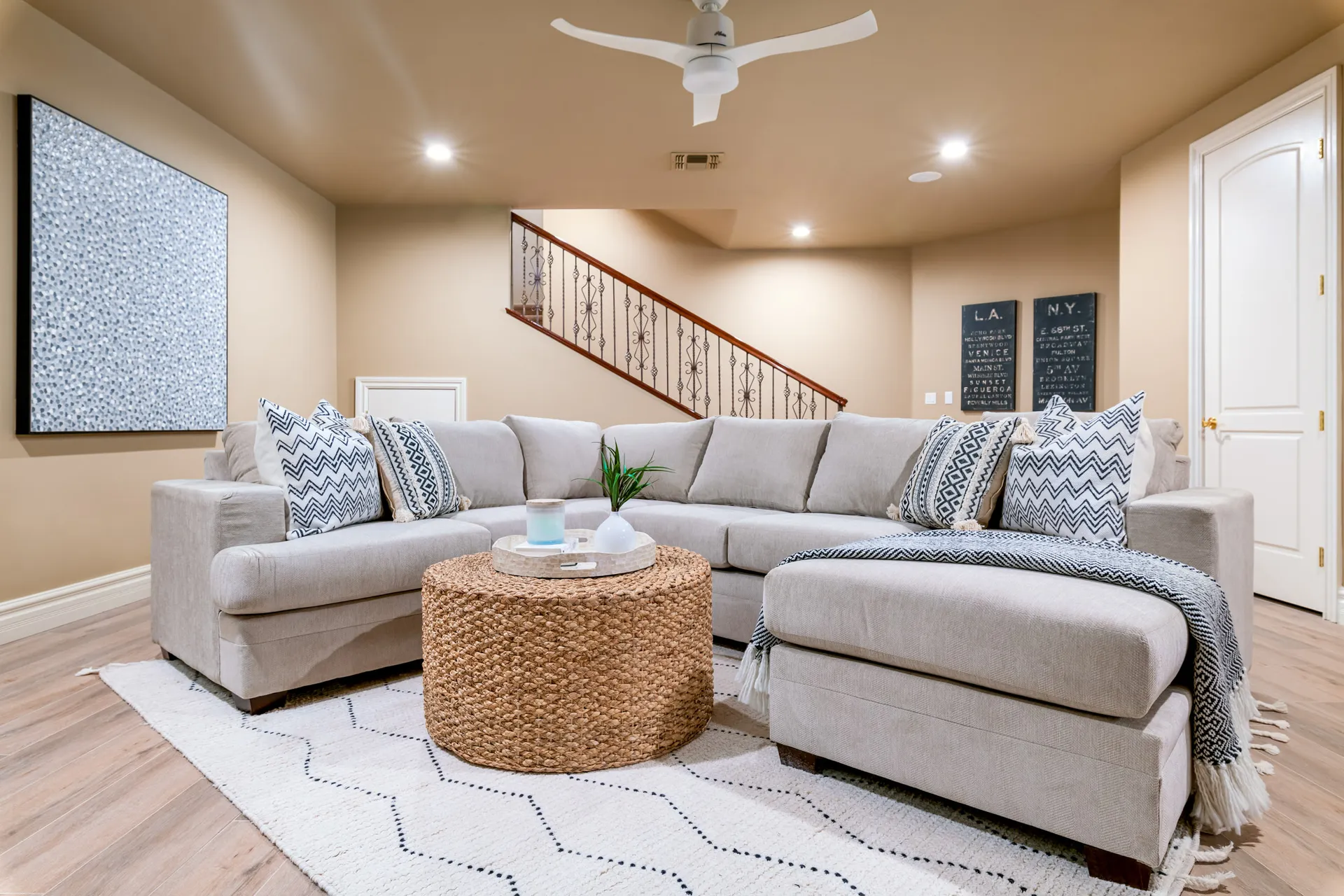 A large L-shaped sectional with patterned pillows, a round woven ottoman, and a metal-railed staircase in a neutral-toned room.