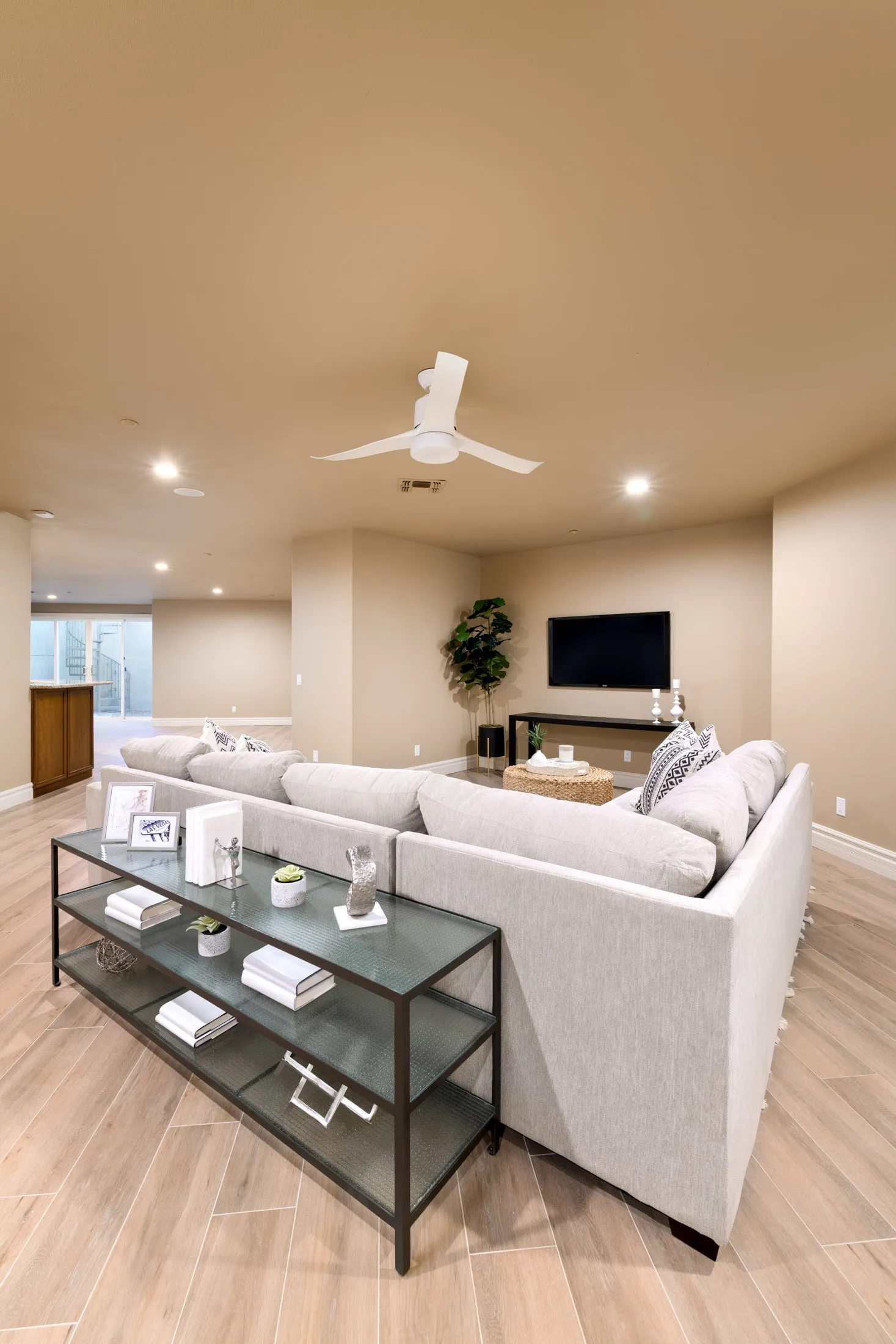A large neutral media lounge with a gray sectional sofa, a glass console table, and a white ceiling fan.