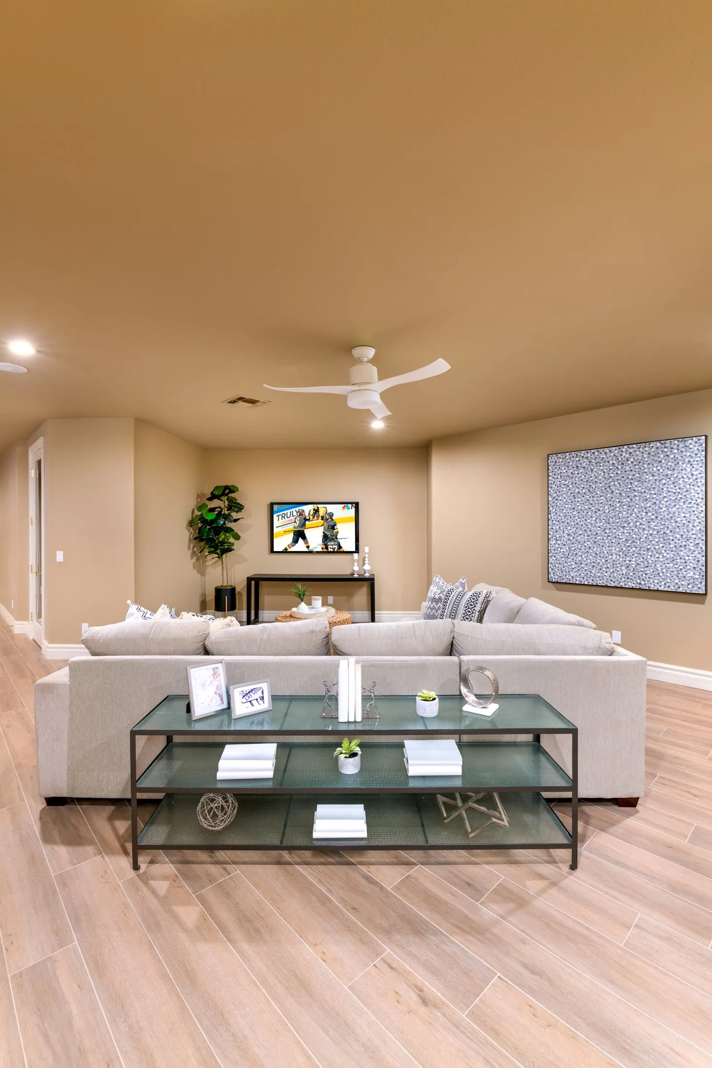 A neutral-toned media lounge with a large sectional, glass console table, and a TV showing a Golden Knights hockey game.