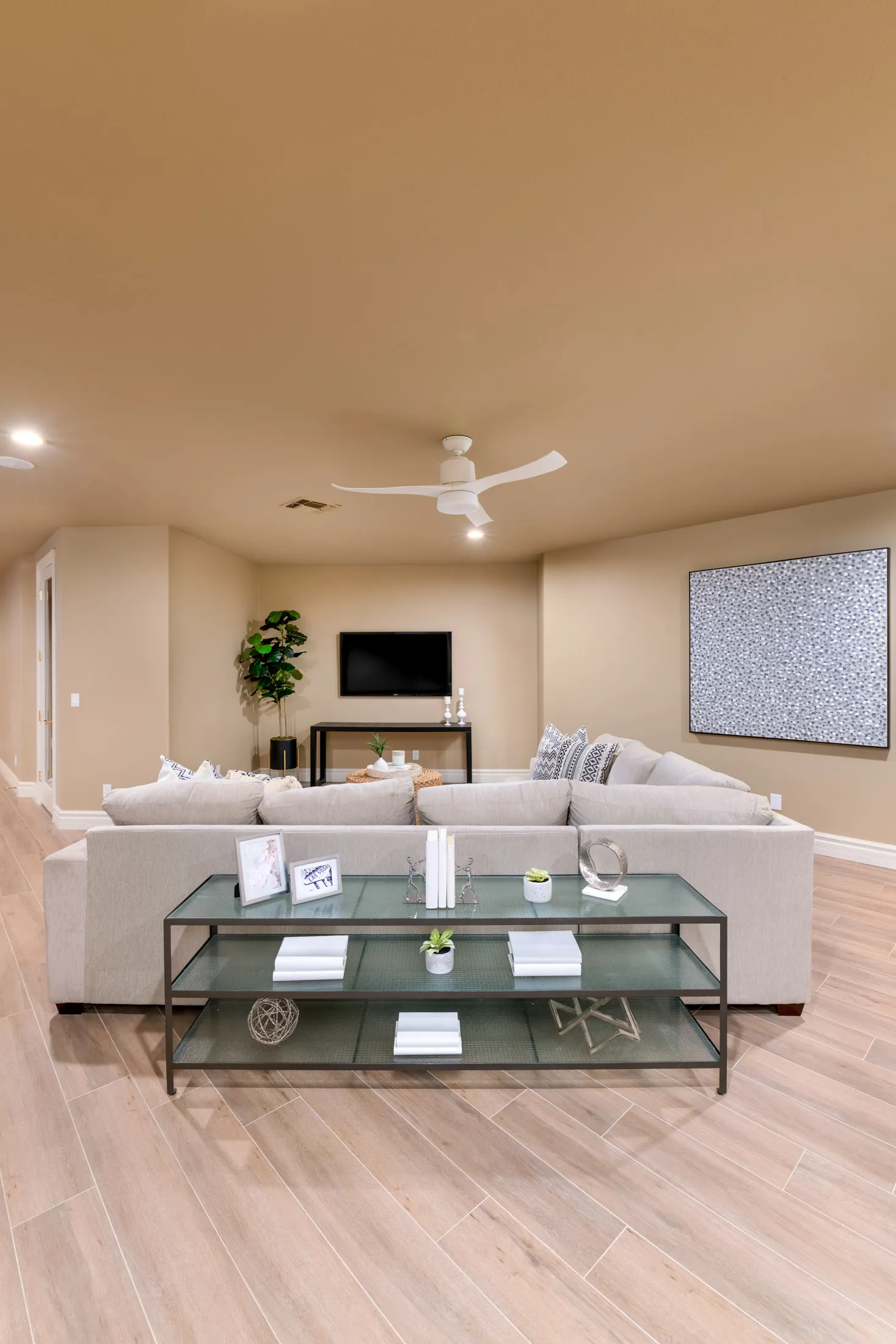 A neutral-toned living area with a large sectional sofa, glass console table, wall-mounted TV, and a white ceiling fan.