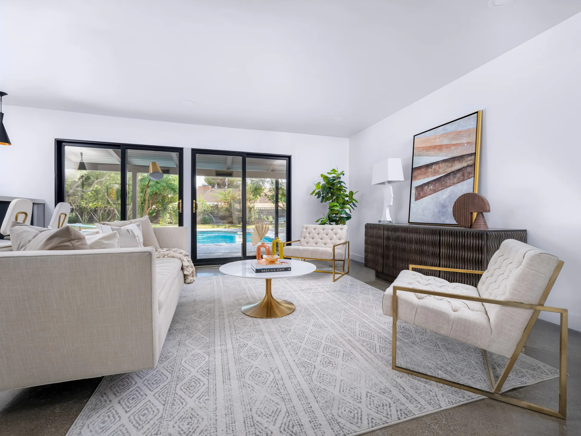 A bright living room with a neutral sofa, tufted chairs framed in gold, and sliding doors that open to a swimming pool view.