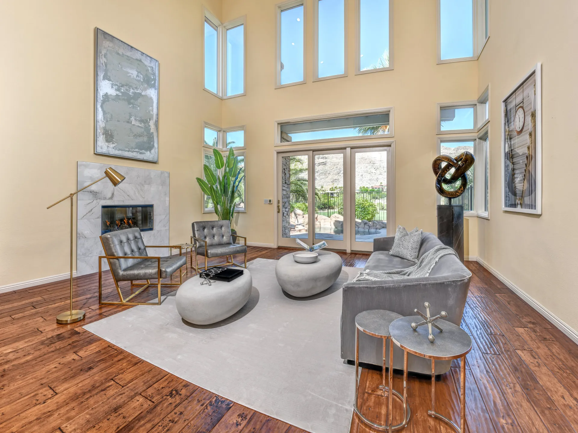 A two-story living area with large windows, a white fireplace, a gray sofa, gold-framed chairs, and sculptural coffee tables.