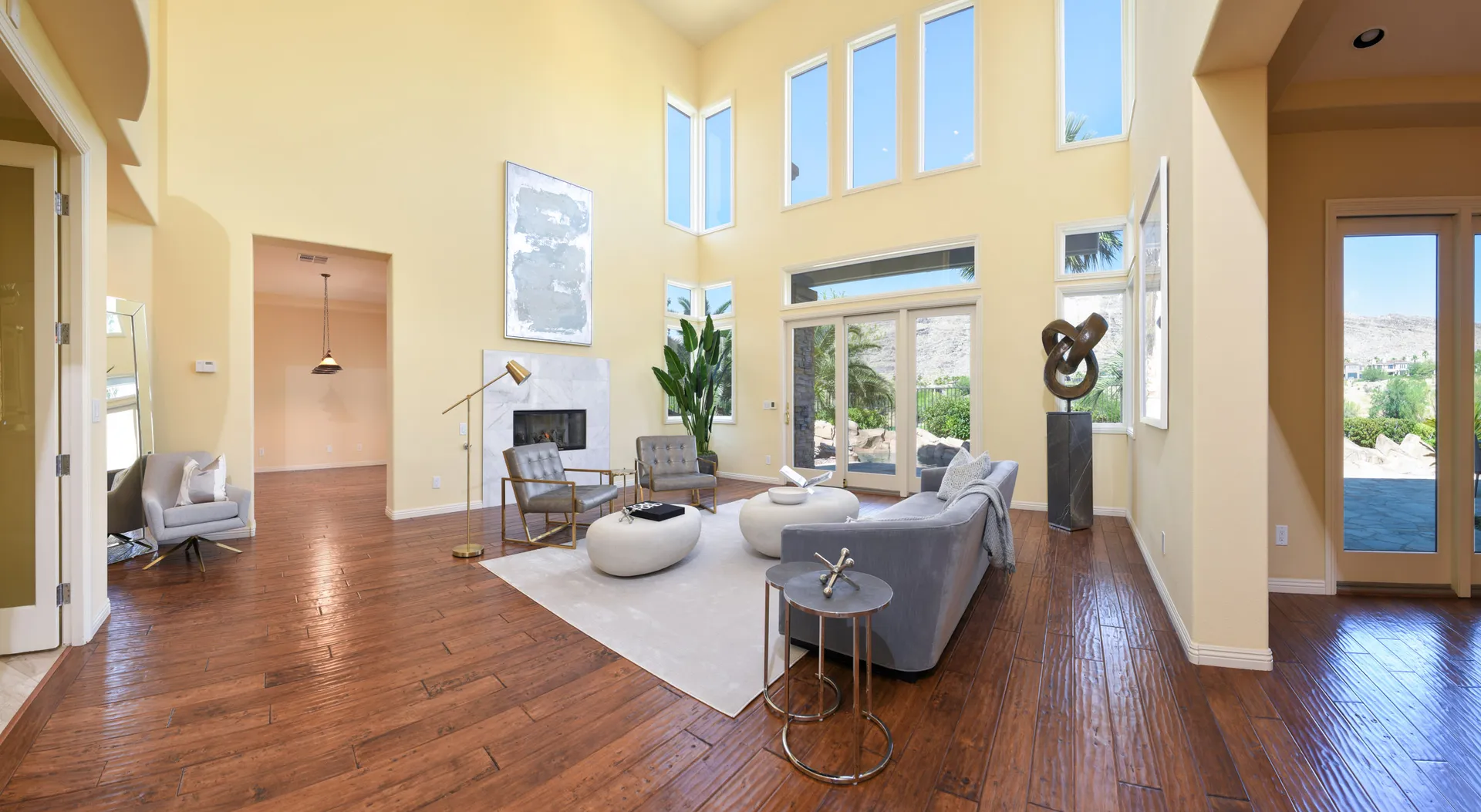A lofty living area with floor-to-ceiling windows, a white fireplace, a gray sofa, and polished wood floors.