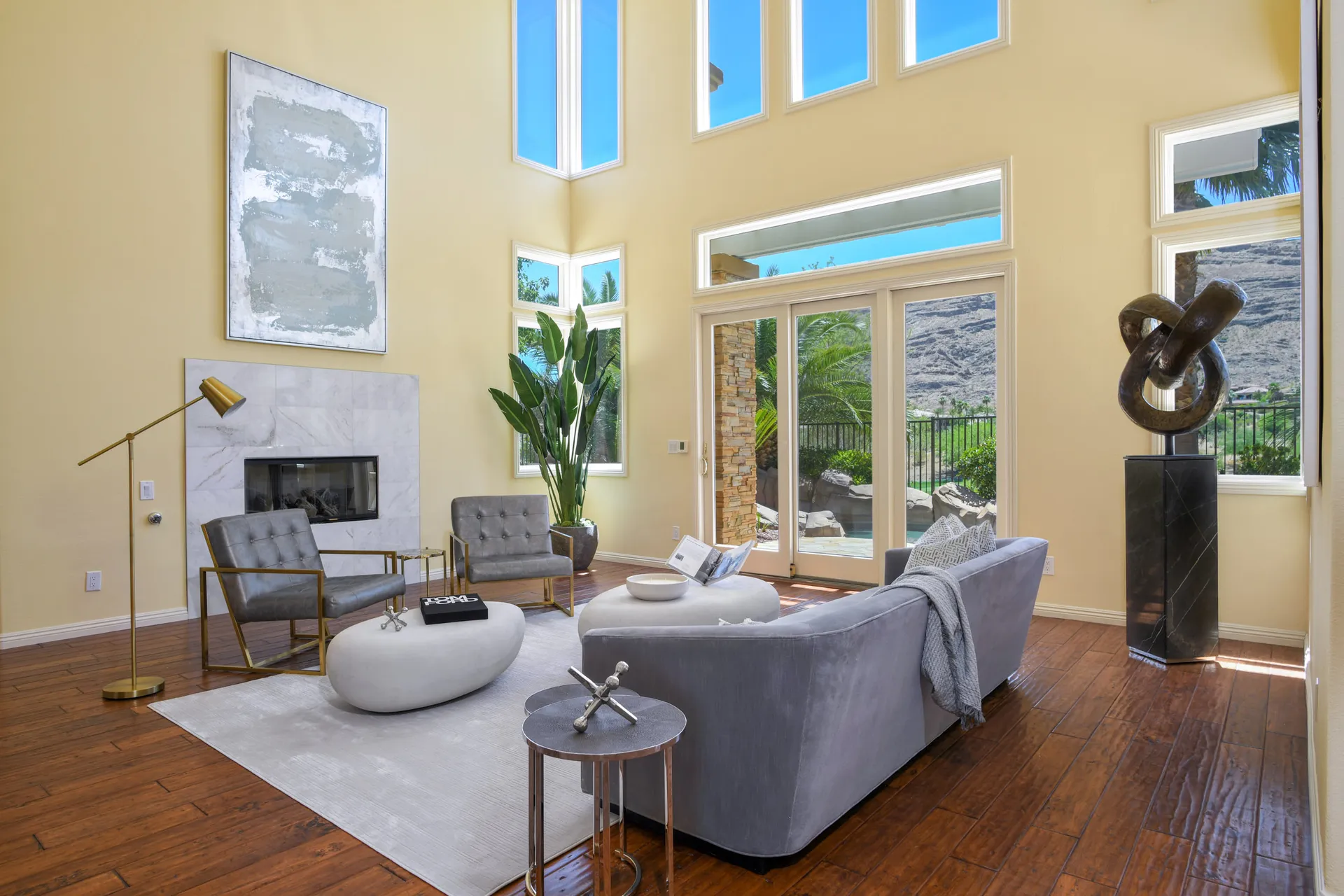 A lofty living room with floor-to-ceiling windows, a white fireplace, and plush gray seating. Large doors lead to an outdoor desert view.