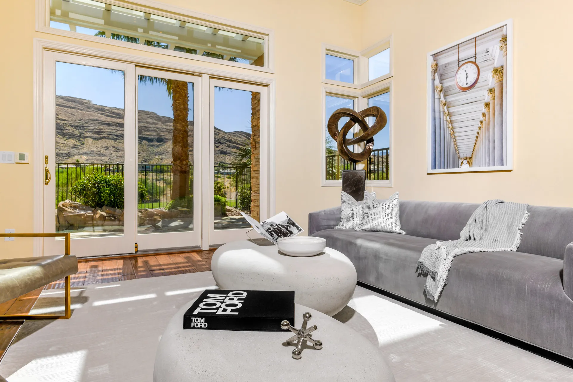 A sunlit living area with a gray velvet sofa, sculptural coffee tables, and large glass doors revealing rocky desert hills.