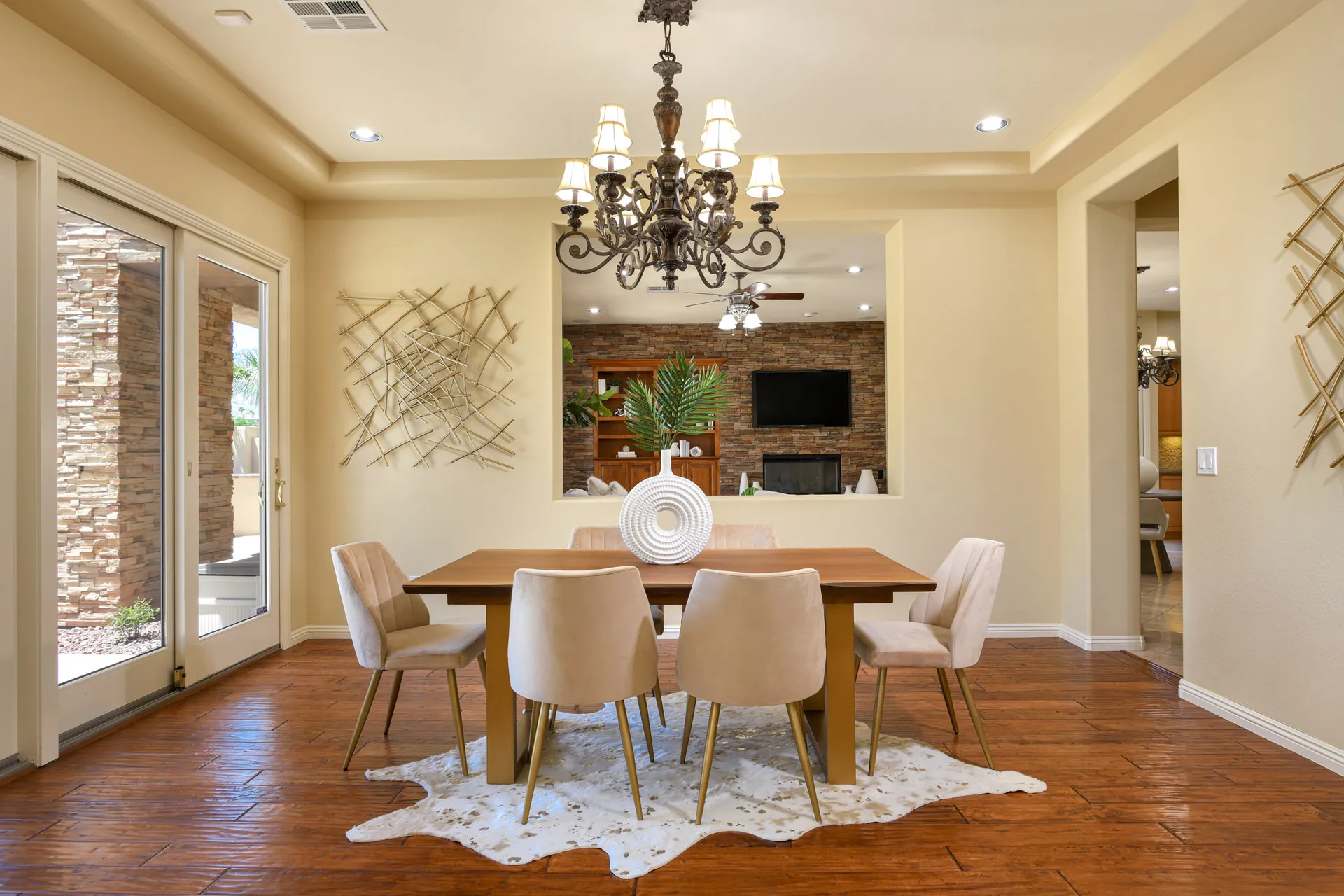 A wooden dining table with plush beige chairs, a white sculptural centerpiece, and a metal wall accent beneath a traditional chandelier.