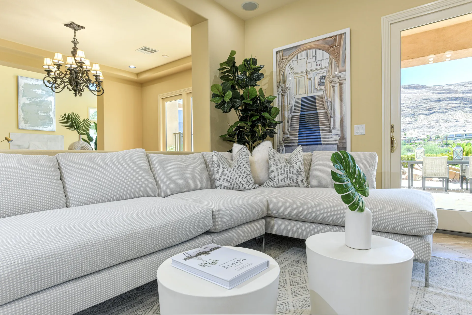 A large light-patterned sectional with decorative pillows, next to a tall plant and a framed photograph of a grand staircase on the wall.