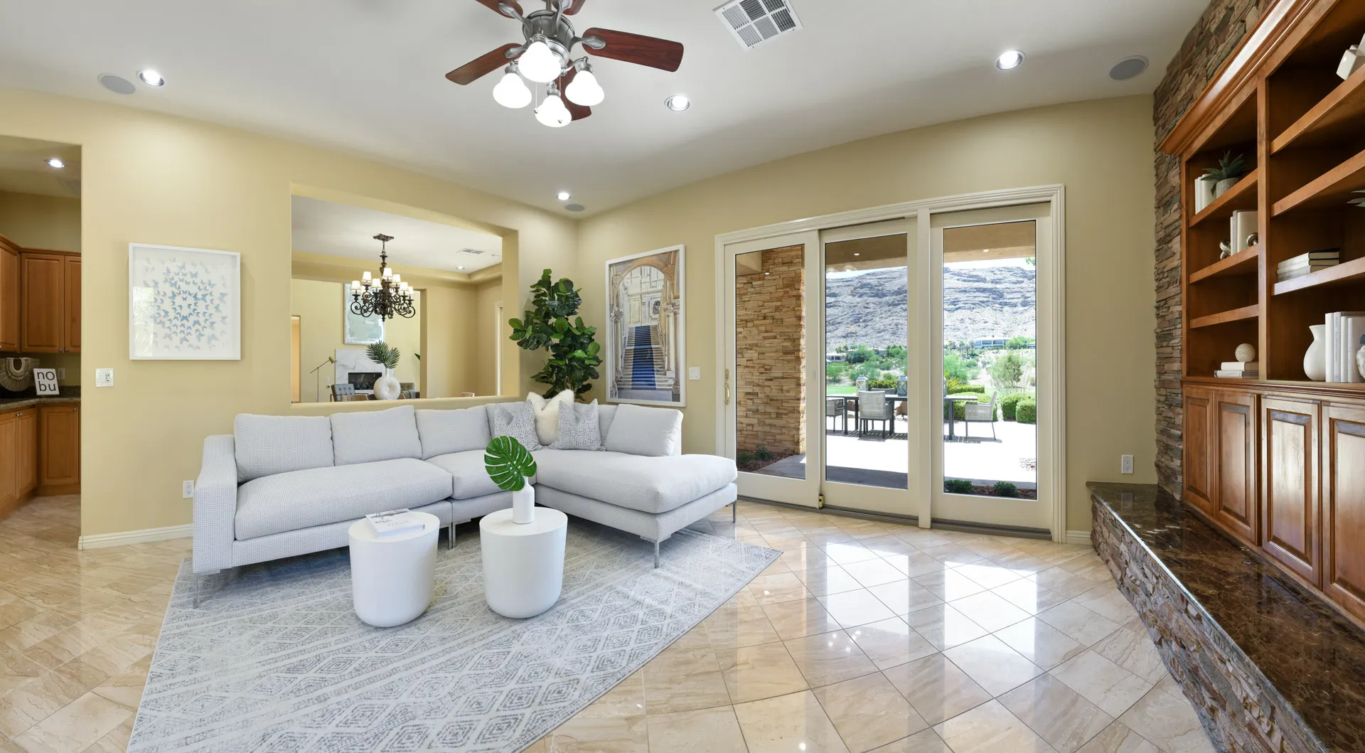 A neutral living area featuring a light sectional, tile floors, a stone-built bookcase, and large doors overlooking a desert landscape.