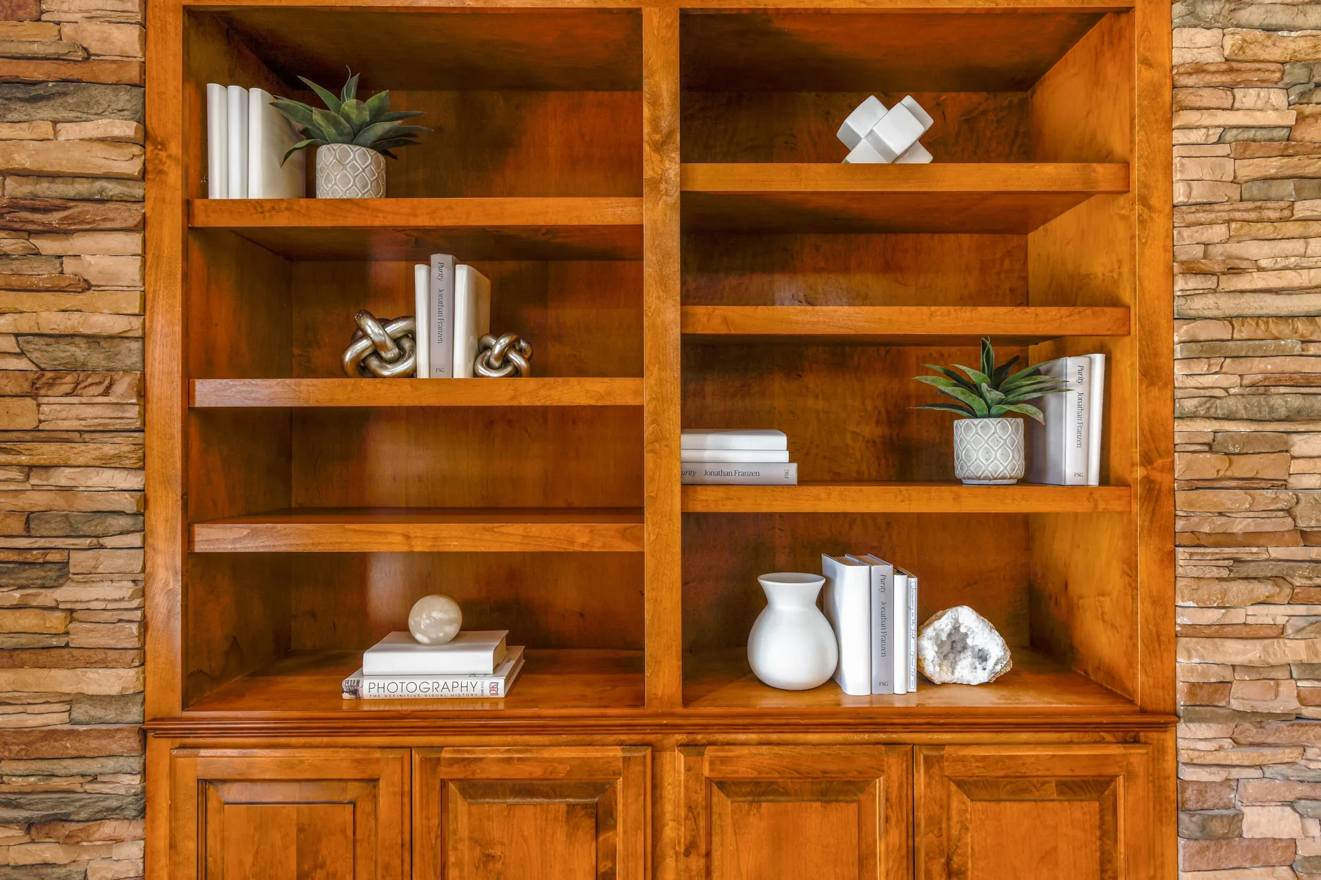A warm wooden bookshelf with decorative items, including potted succulents, white books, and sculptural accents.
