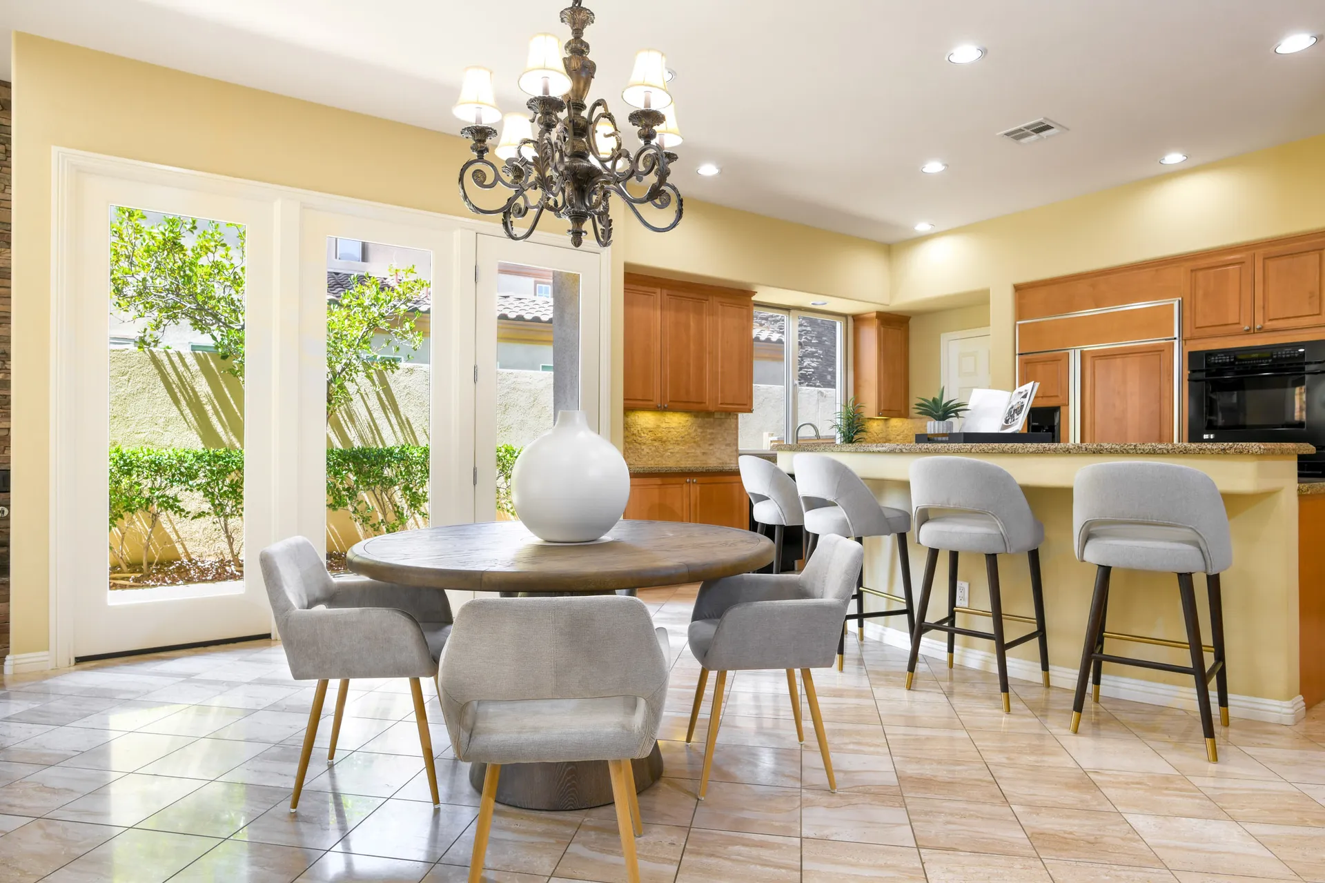 A round wooden dining table with a white vase, surrounded by gray chairs, next to a granite kitchen island with matching barstools.