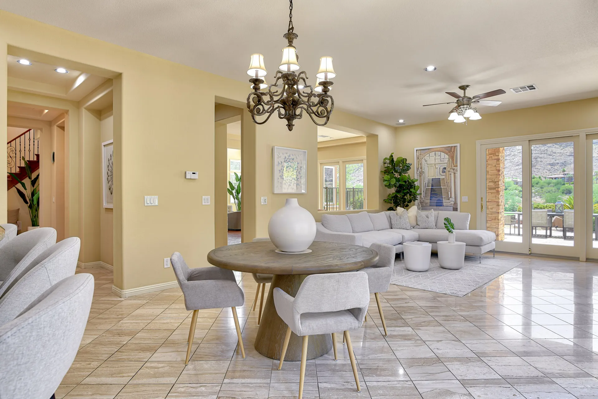 A round wooden dining table with a white vase, surrounded by gray chairs, adjacent to a light sectional sofa in a warm, open-concept space.