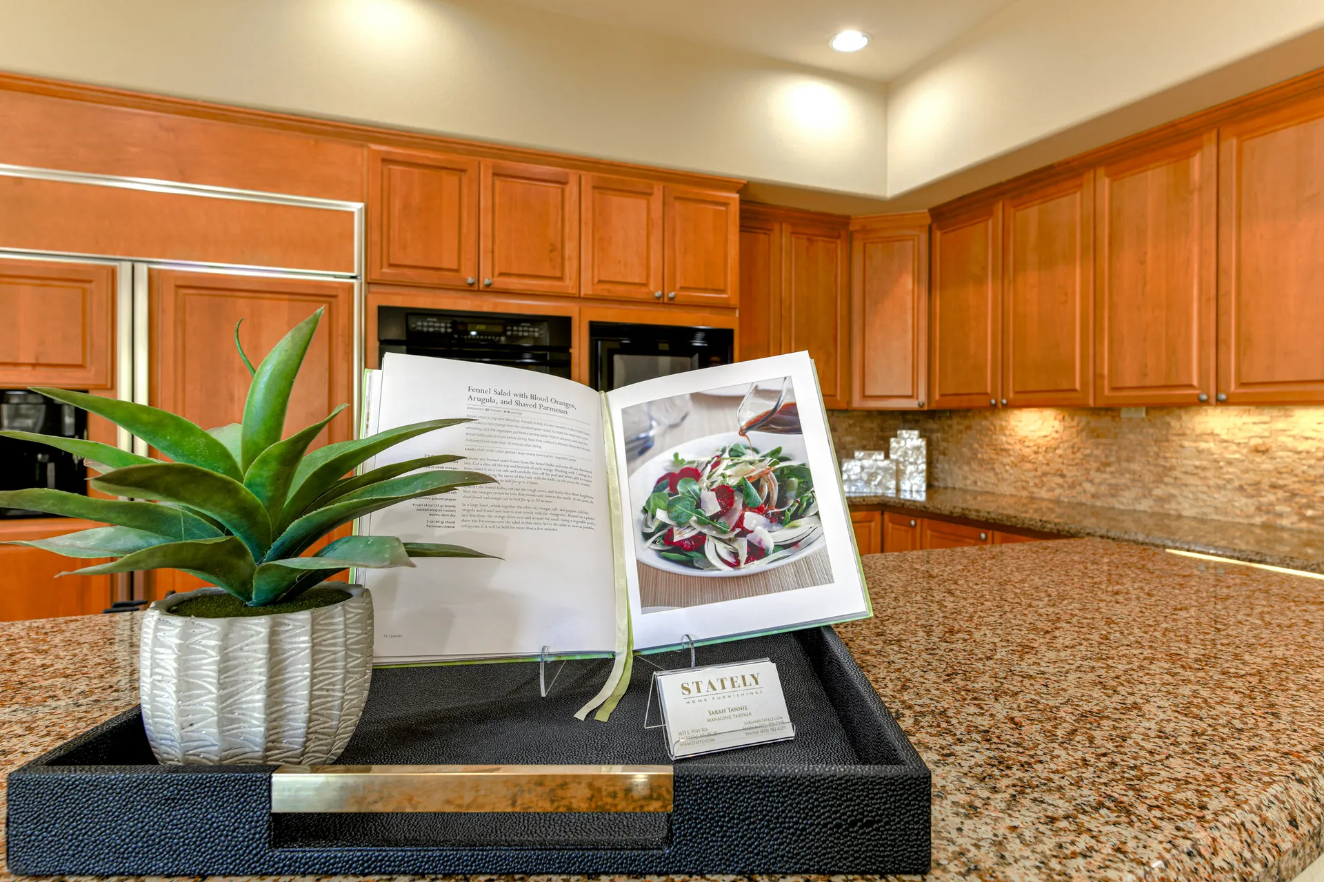 A warm wood kitchen with a granite countertop, featuring an open cookbook and a small succulent in a decorative pot.