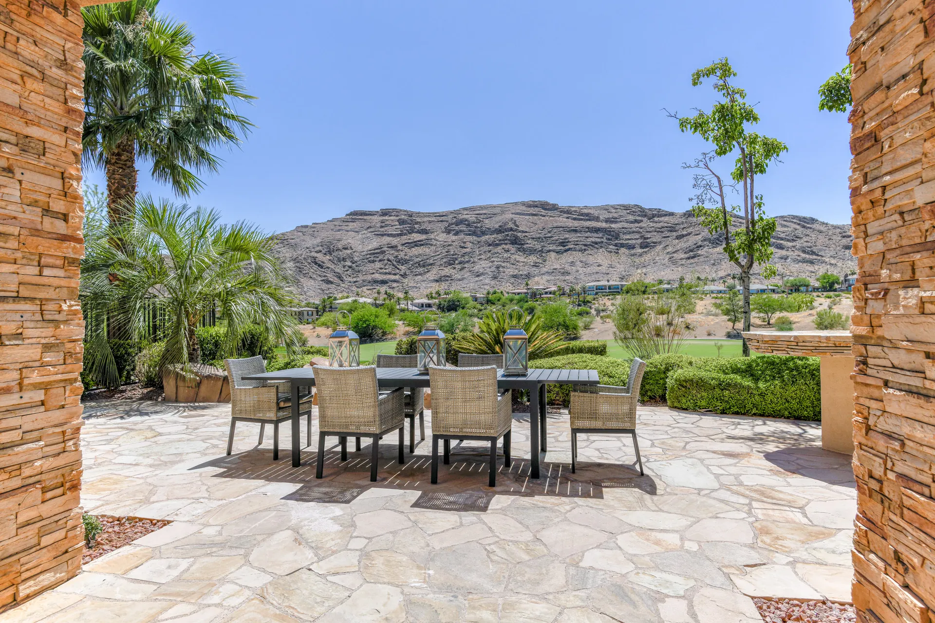 A large black outdoor dining table with woven chairs on a stone patio, framed by desert landscaping and rocky hills in the background.