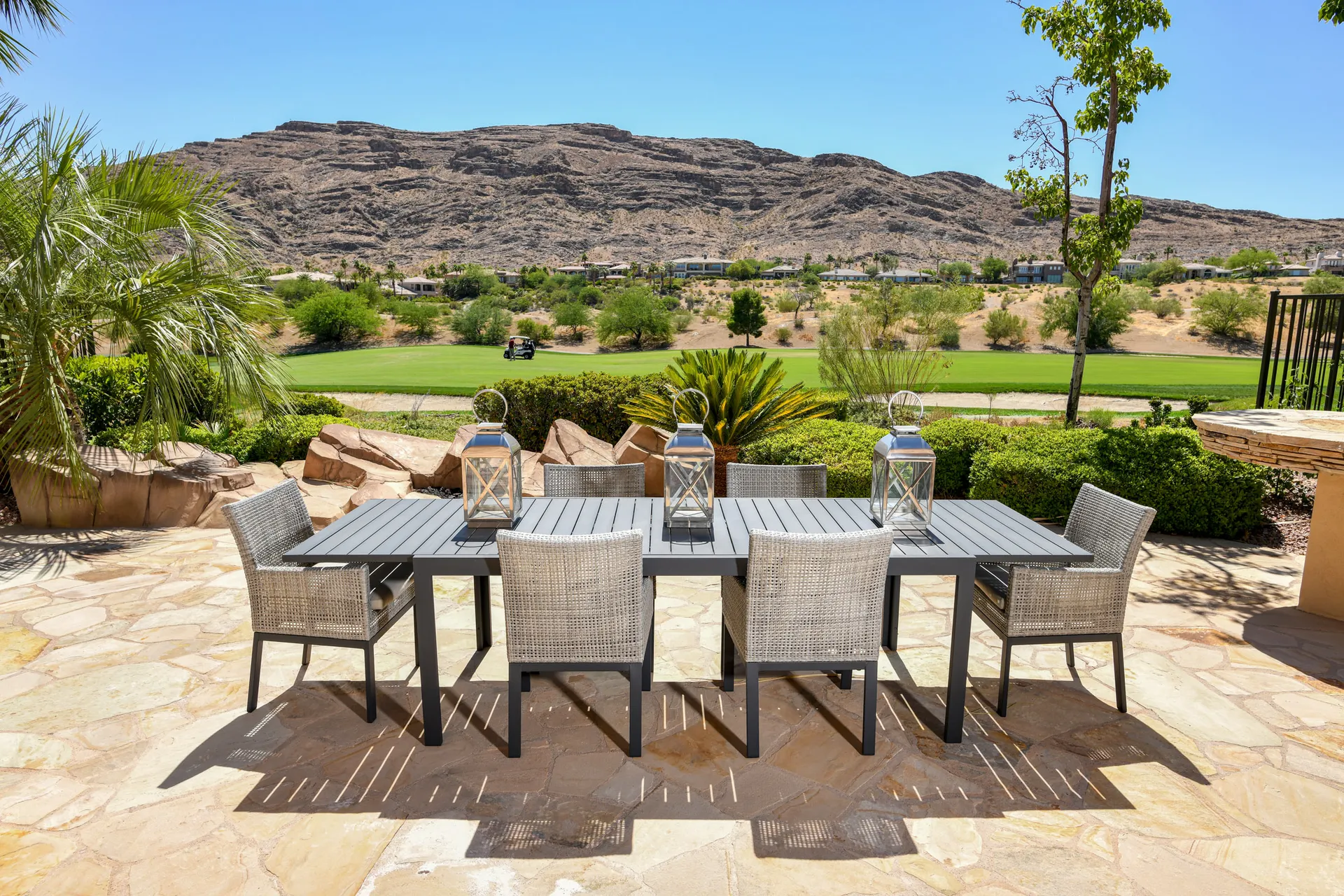 A spacious stone patio with a rectangular black dining table, wicker chairs, and lanterns, overlooking a golf course and rocky hills.